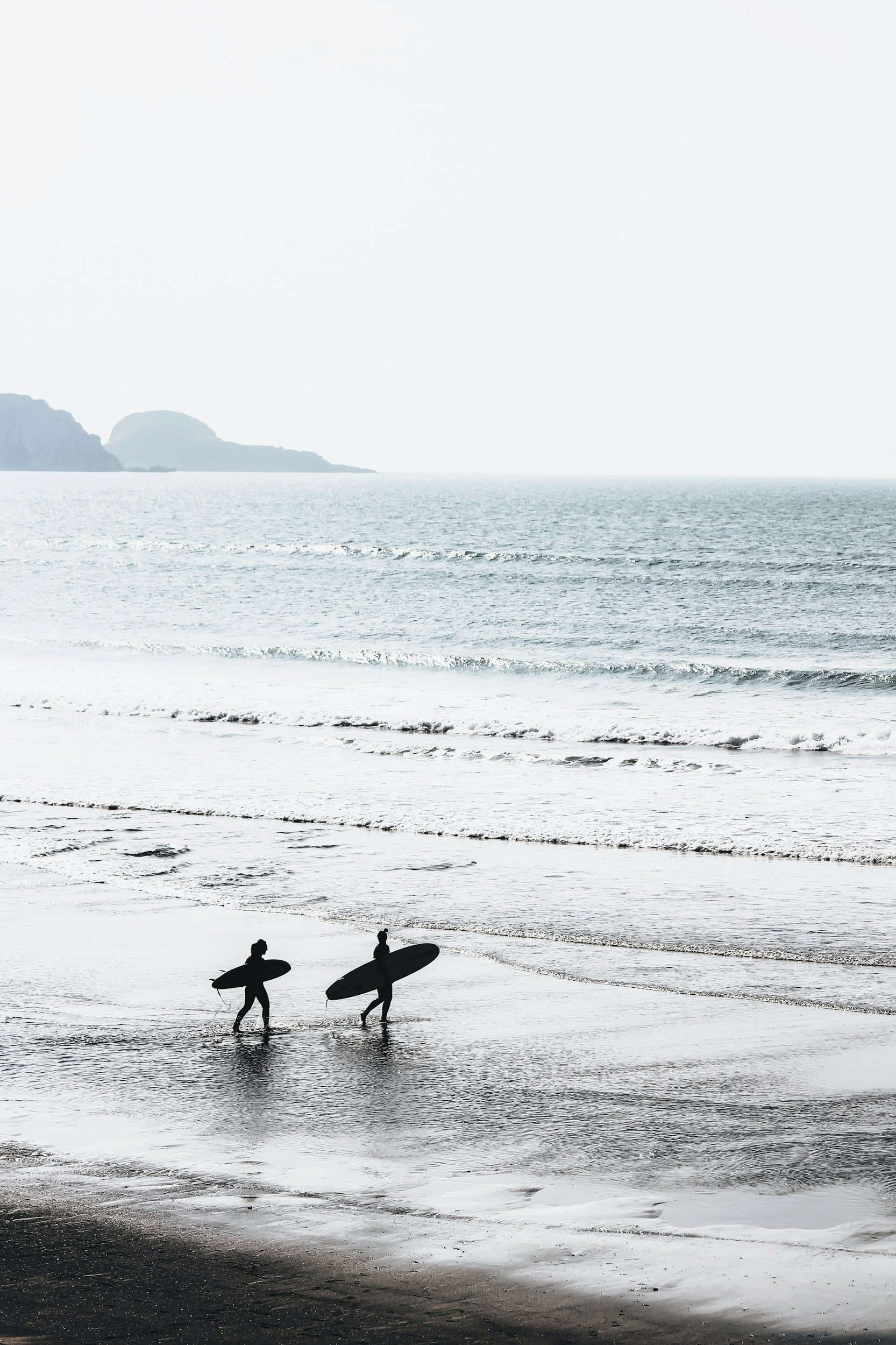 Two surfers walking into the ocean with surfboards on a beach during day with calm waves and distant mountains.