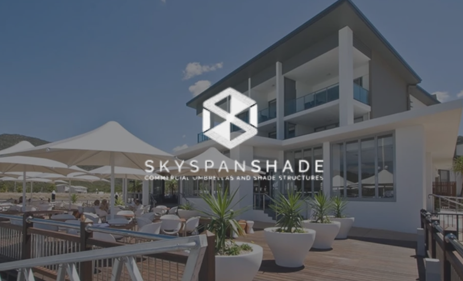 A modern multi-story building with outdoor seating area and large umbrellas, featuring the logo and text for Skyshade, specializing in commercial umbrellas and shade structures.