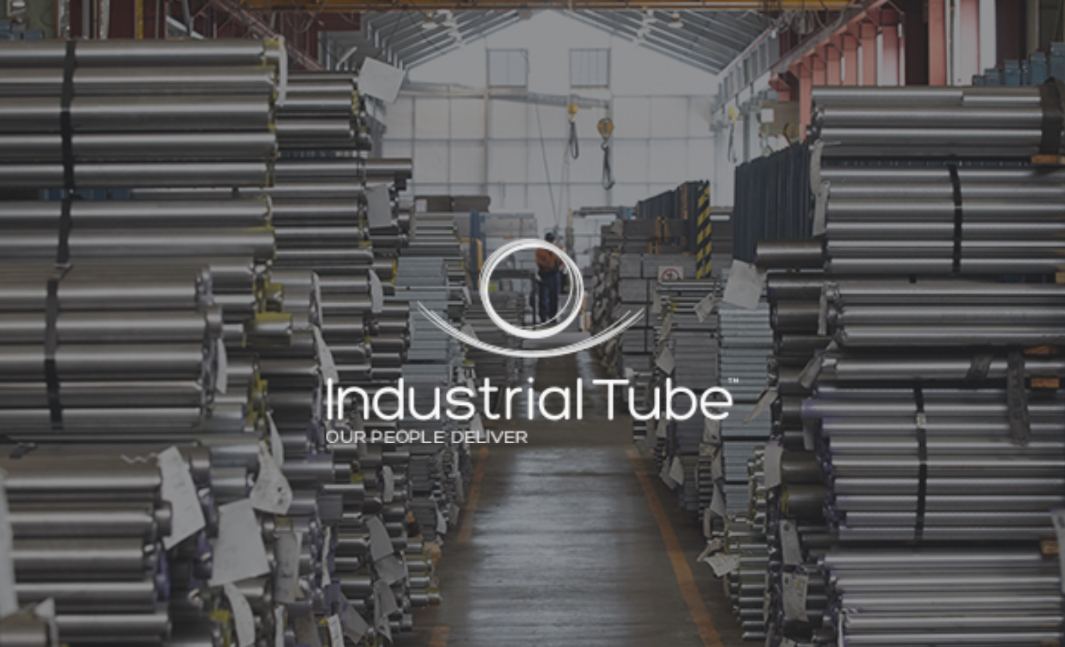 Industrial warehouse with stacked metal pipes on both sides and a worker walking in the background, with the overlay text 'Industrial Tube, Our People Deliver'.