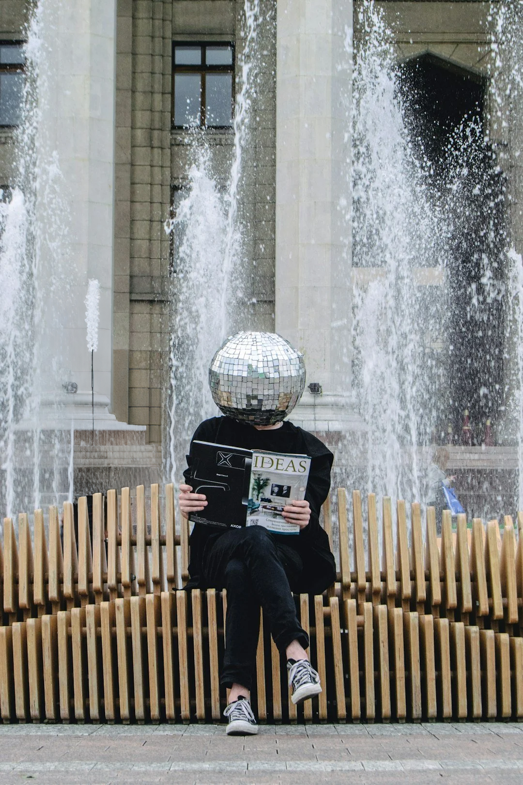 Person sitting on a wooden bench in front of a fountain with a globe-shaped mirrored head, reading a magazine titled 'IDEAS'.
