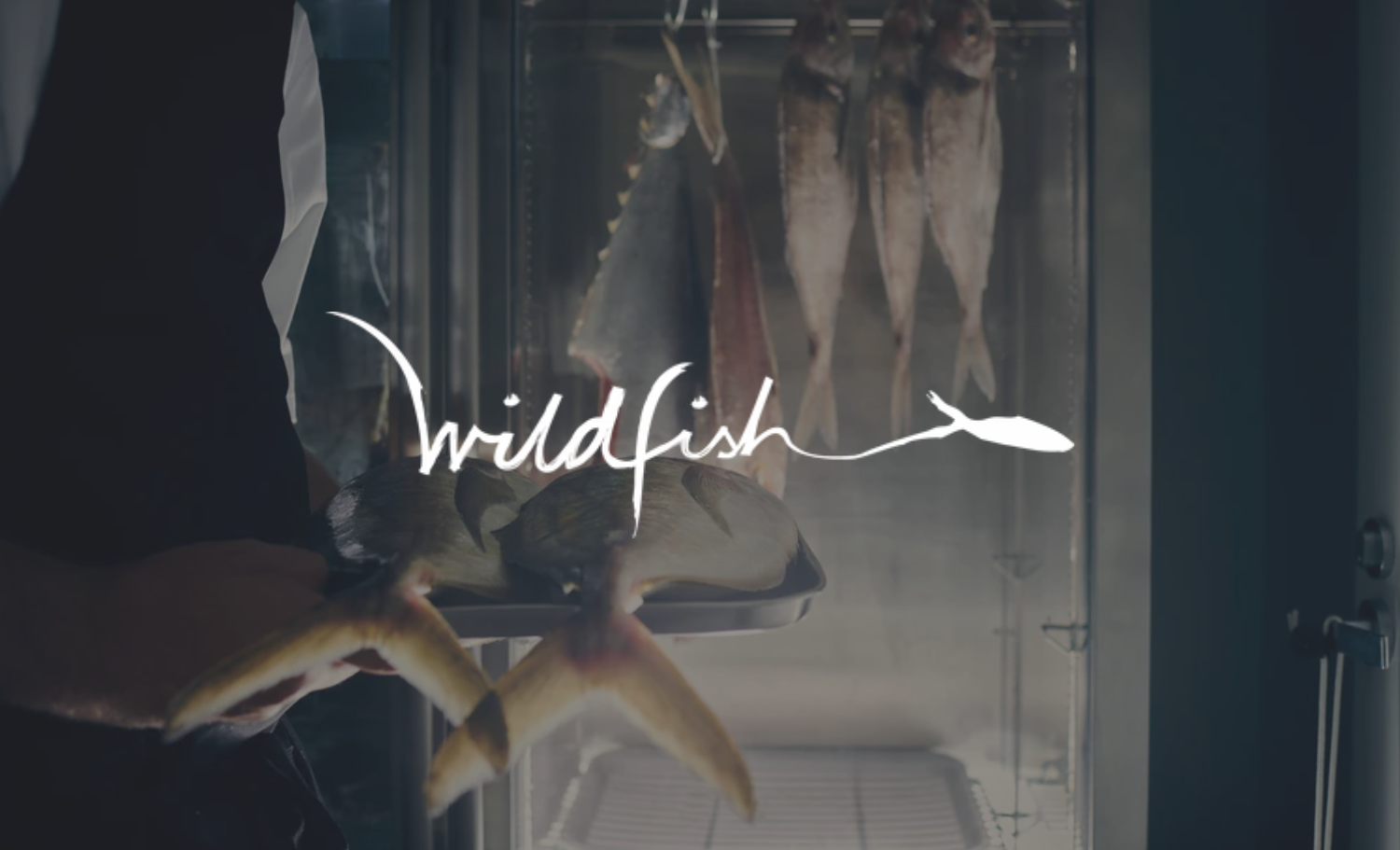 A person holding a tray with fish in front of a glass case with hanging fish inside a fish market or restaurant.