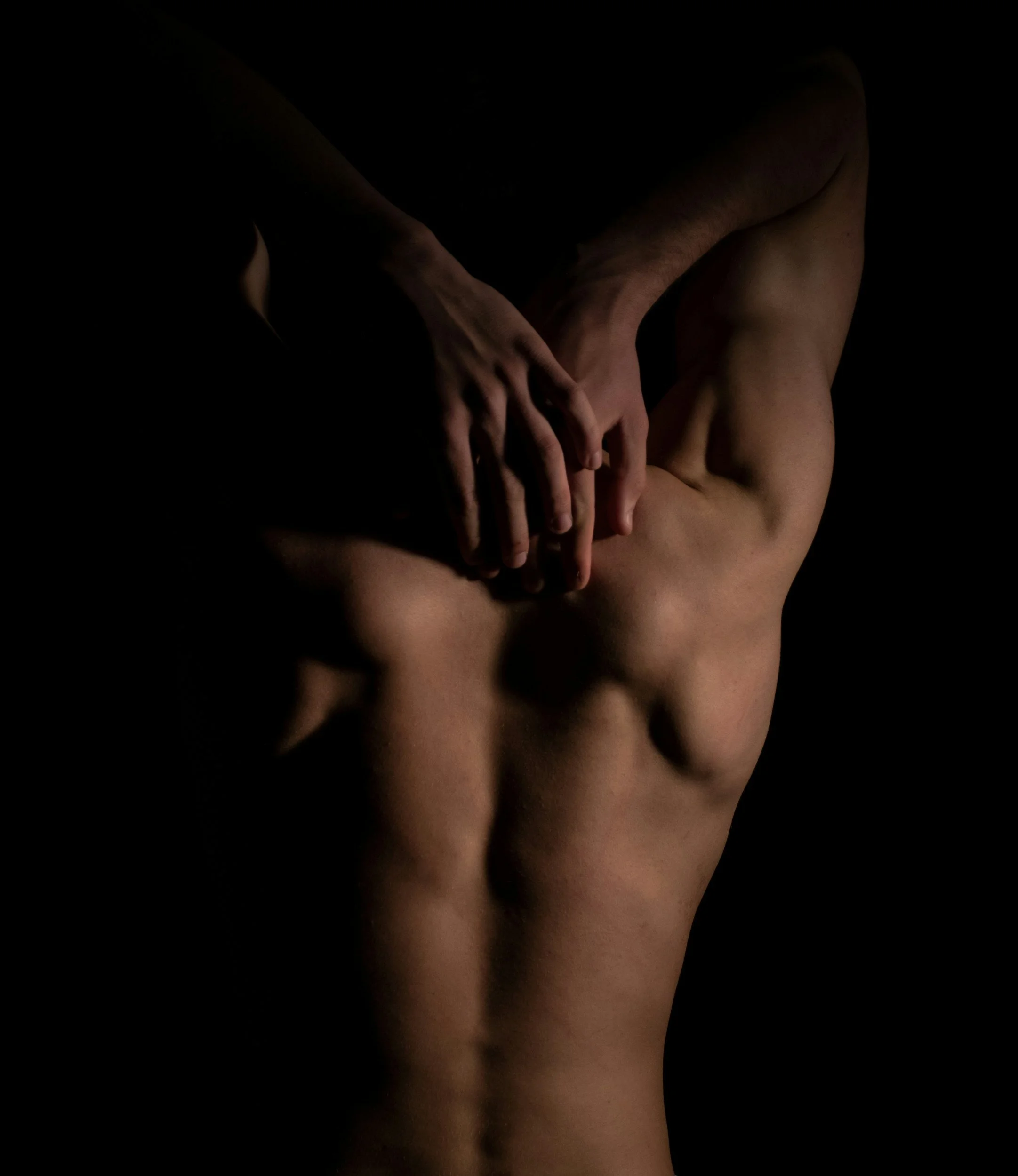 A person's bare back and shoulder with another person's hands placed on their upper back in a dark setting with focused lighting.