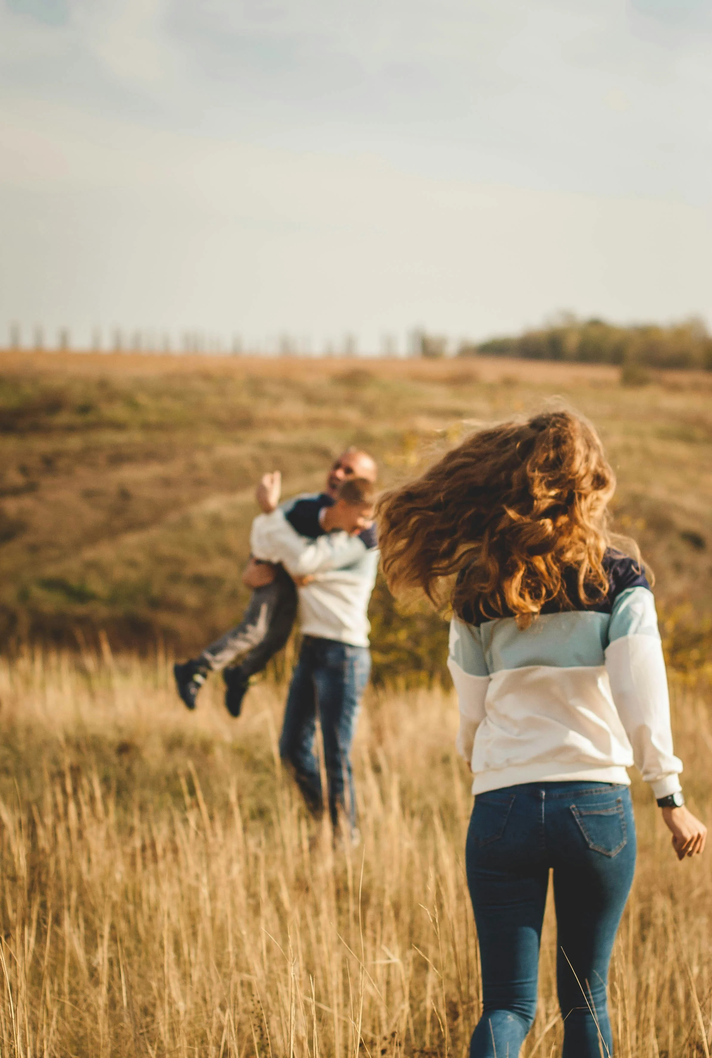Family playing in a grassy field during sunset, a woman stands in the foreground with her back to the camera, and a man is lifting a child in the background.