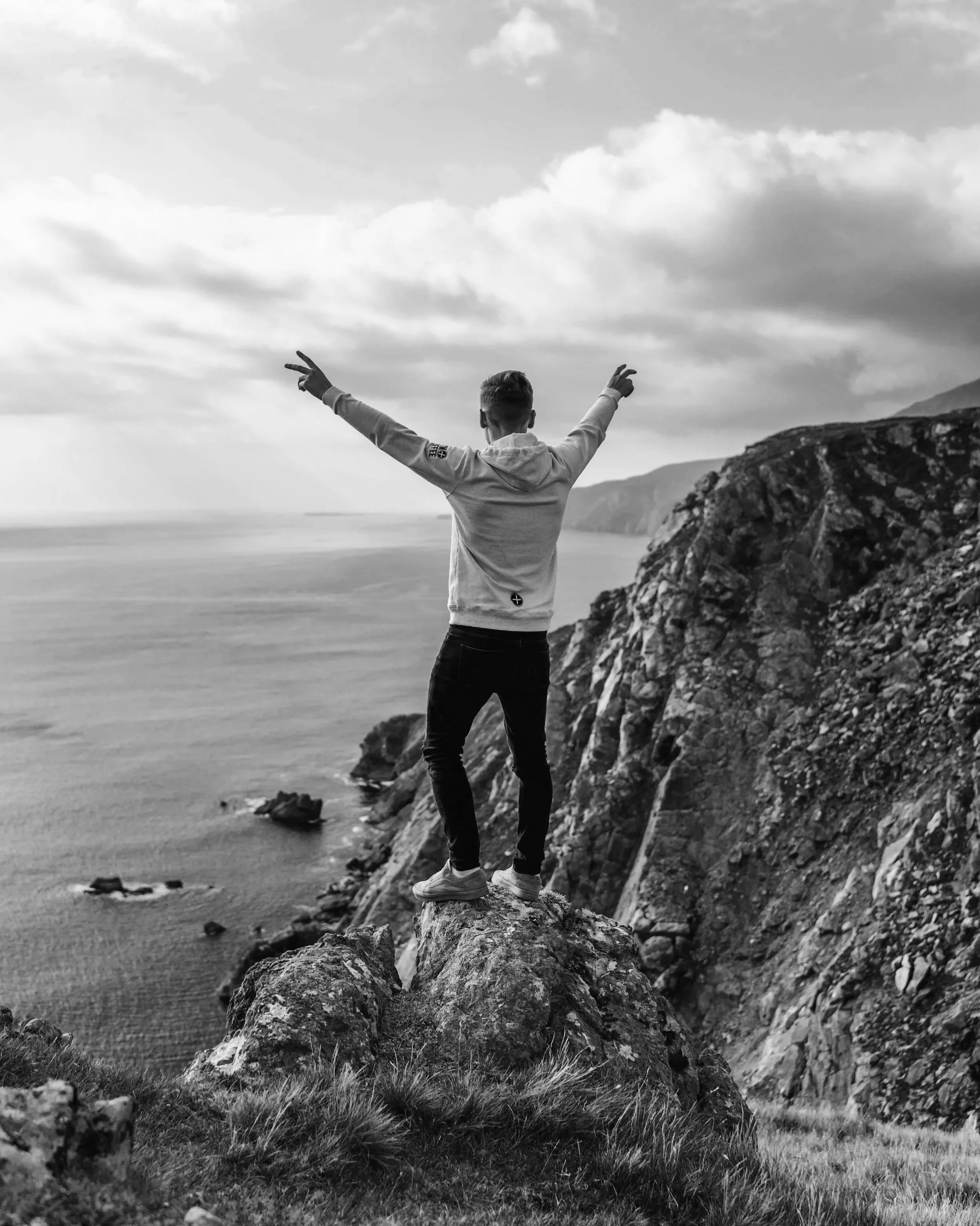 A person standing on a rock ledge with arms outstretched, overlooking a coastal landscape with cliffs and ocean in black and white.