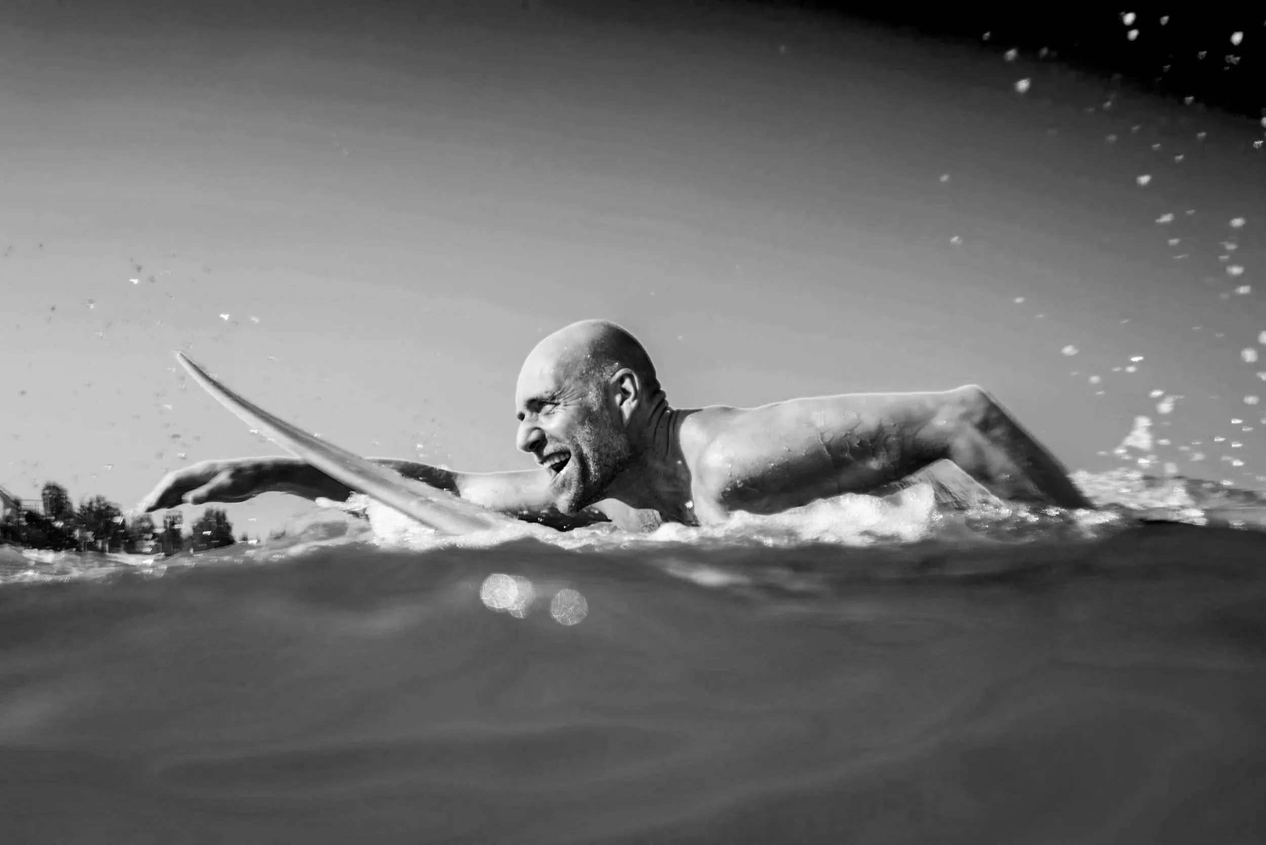 A black and white photograph of a man swimming freestyle in open water, appearing determined and focused with a bald head and strapless swimsuit. Man living an active, energized life — representing the daily habit of Atlas Performance Pro