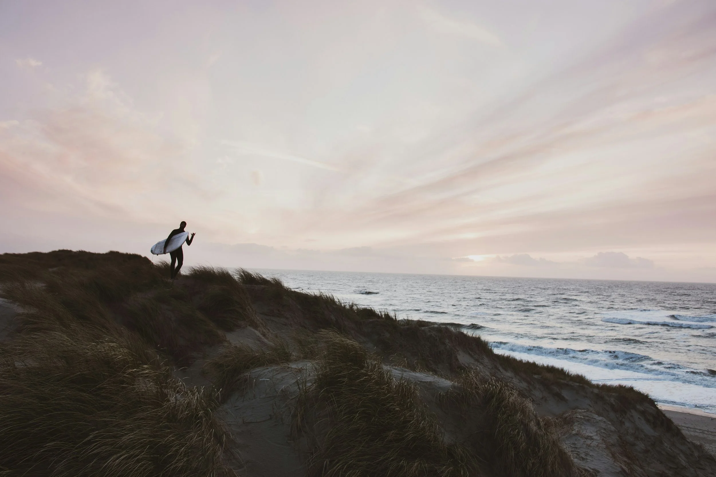 A person with a surfboard walking along a sandy dune overlooking the ocean at sunset.
