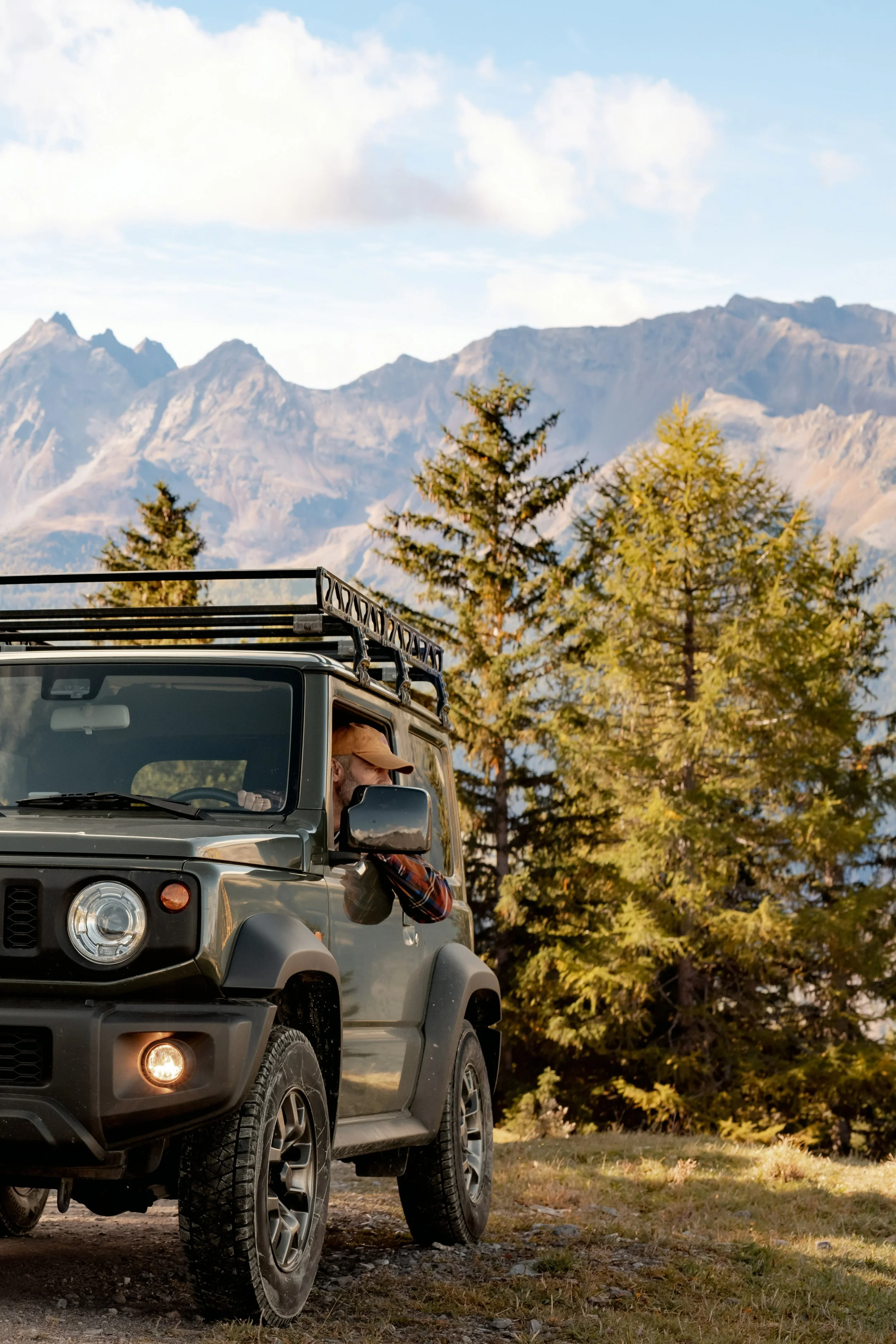 A person looking out of the driver side window of a parked black off-road vehicle, with trees and mountain peaks in the background on a clear day.