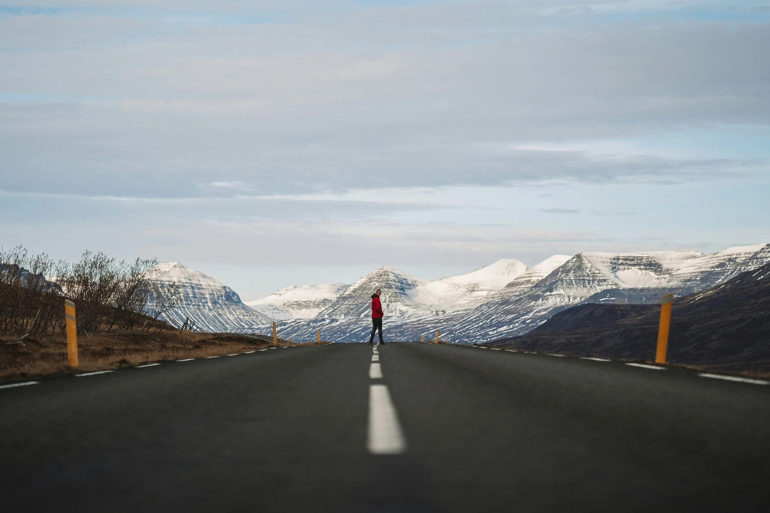 A person in a red jacket standing alone in the middle of a long, empty road surrounded by snow-capped mountains and a cloudy sky.