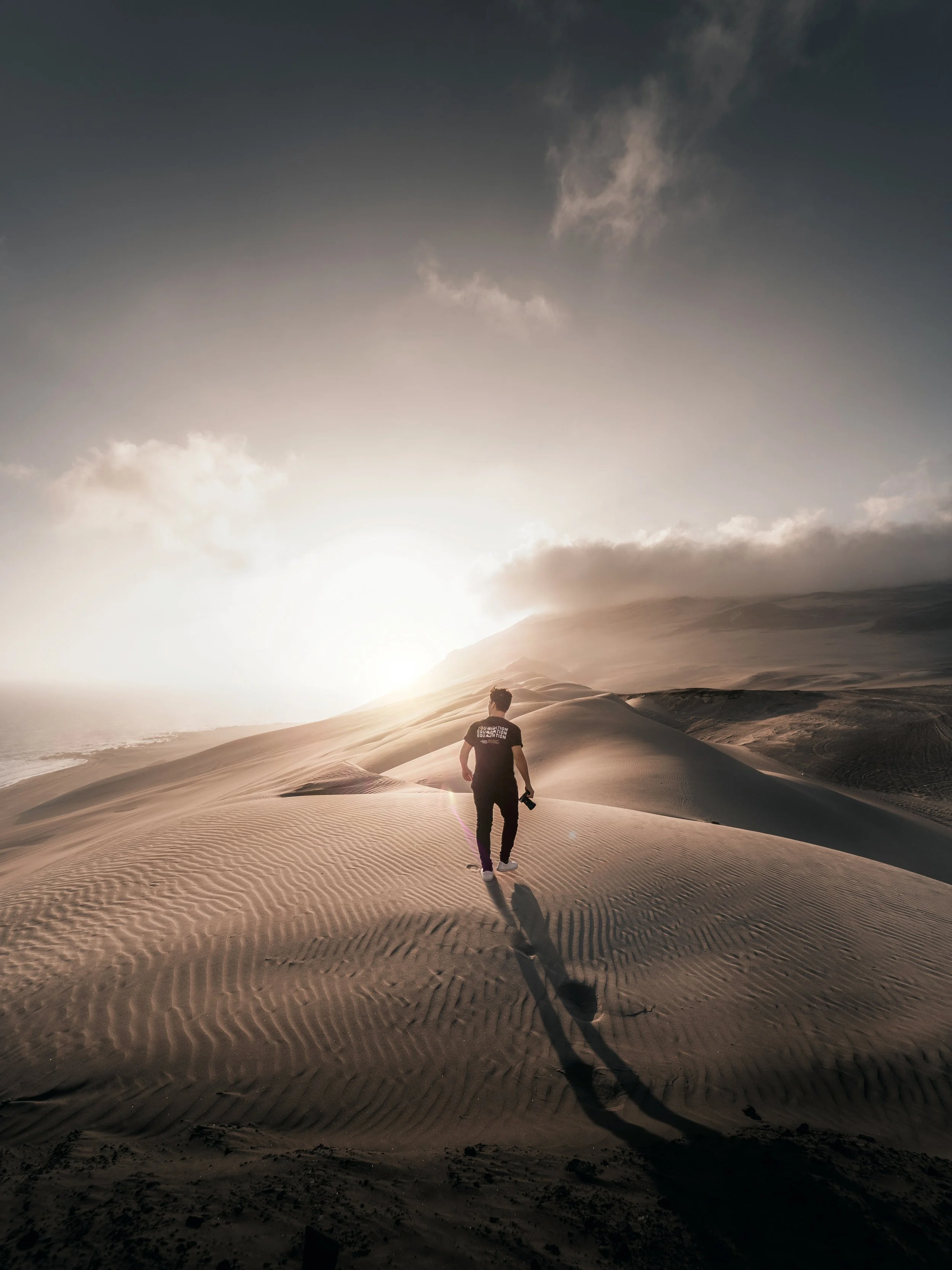 Person walking on sand dunes at sunset or sunrise with clouds in the sky.
