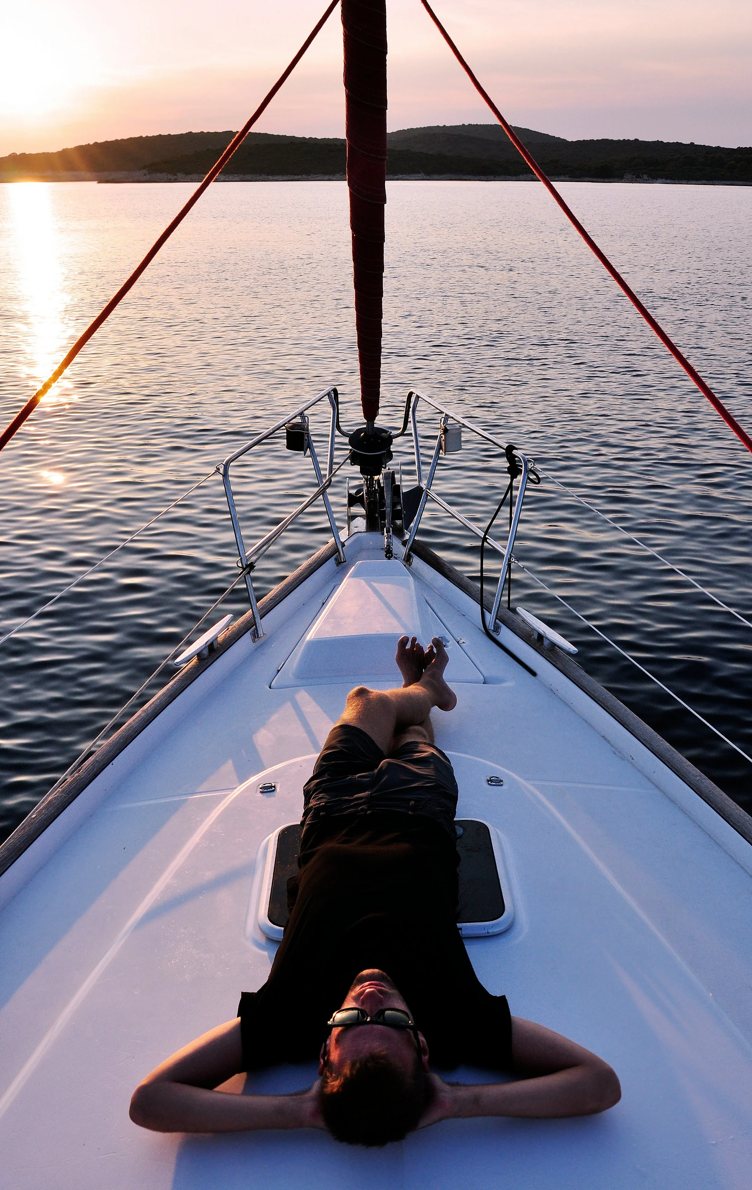 A person lying on the deck of a sailboat during sunset, with legs crossed, wearing sunglasses and a black shirt, surrounded by water and distant hills.
