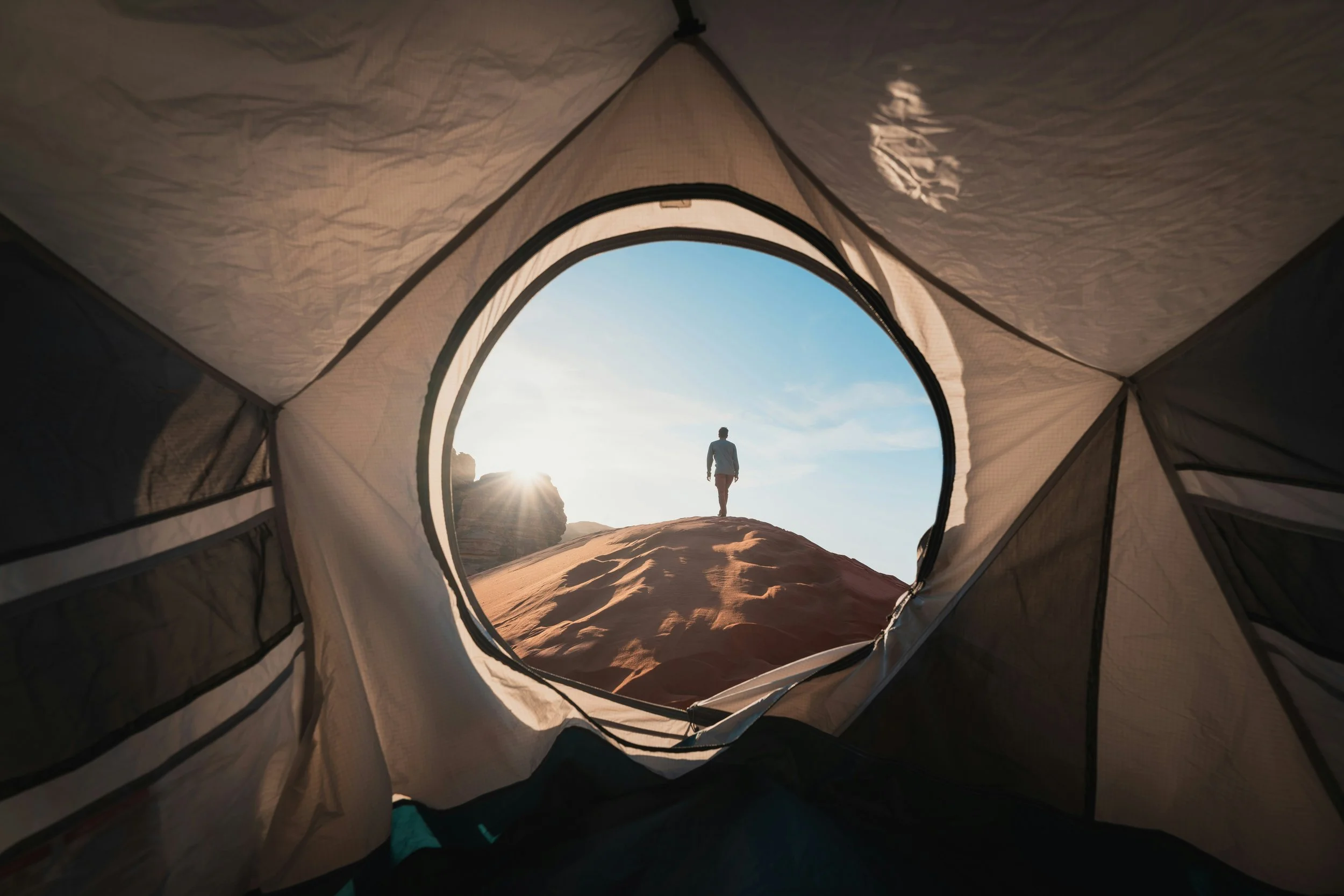 View from inside a tent looking out at a person walking on a sandy hill at sunset or sunrise with blue sky and clouds in the background.