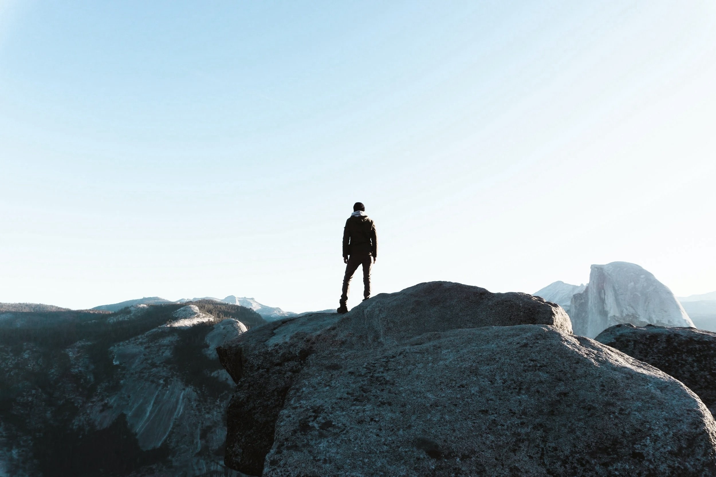 A person standing on a large rock overlooking a mountainous landscape with clear sky.