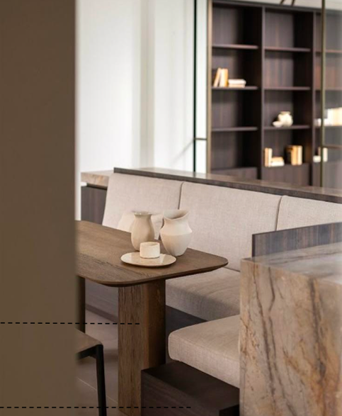 Interior of a modern living space with a beige upholstered bench, wooden table with pottery, and a dark bookshelf with decorative items.