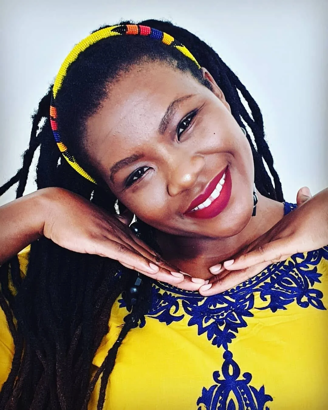 Smiling woman with dark dreadlocks wearing a yellow dress with blue embroidery, a colorful beaded headband, and red lipstick, posing with hands under her chin.
