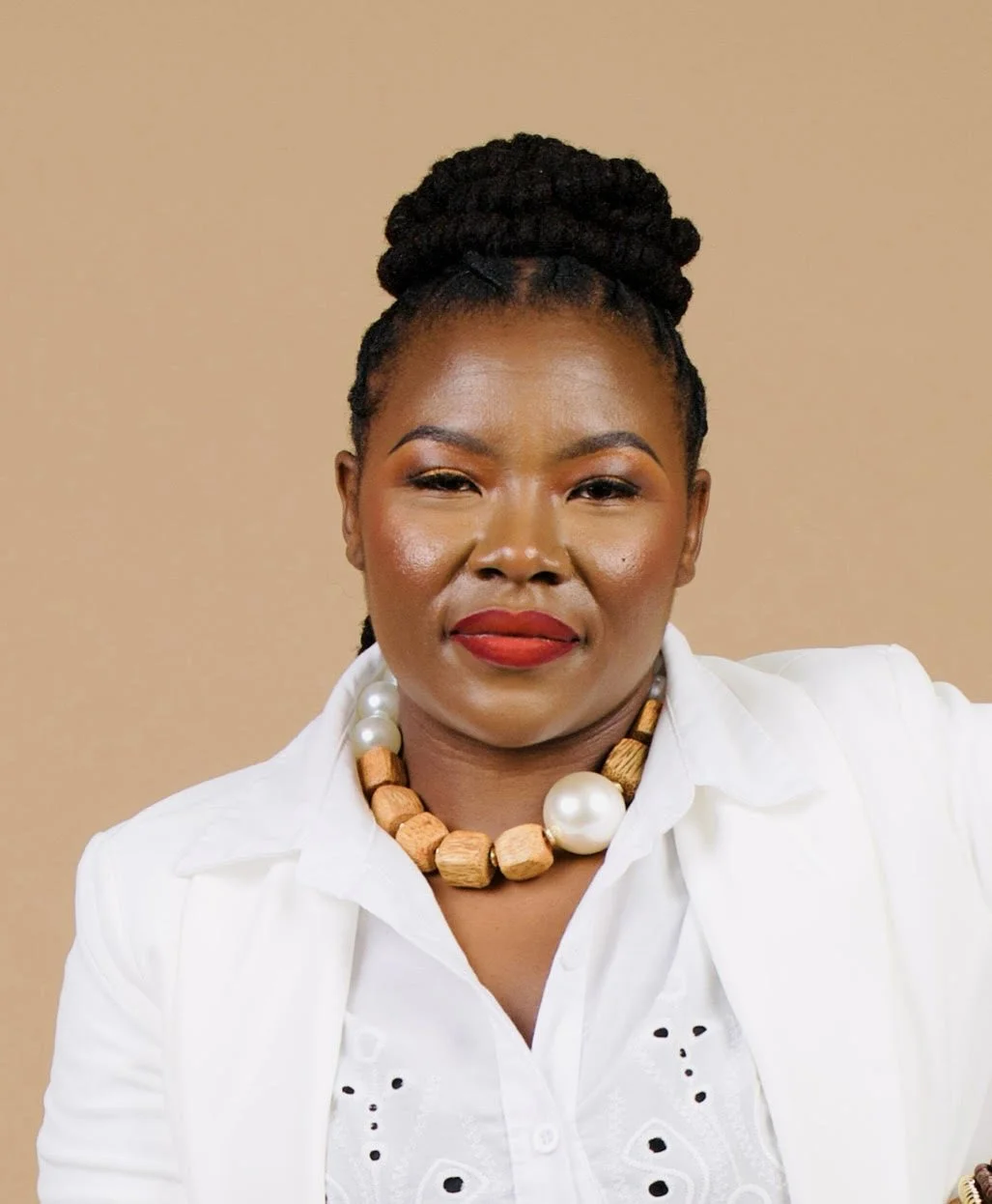A woman with dark skin and braided hair styled in an updo, wearing a white shirt with embroidered details, a chunky wood and pearl necklace, and bright red lipstick, posing against a plain beige background.