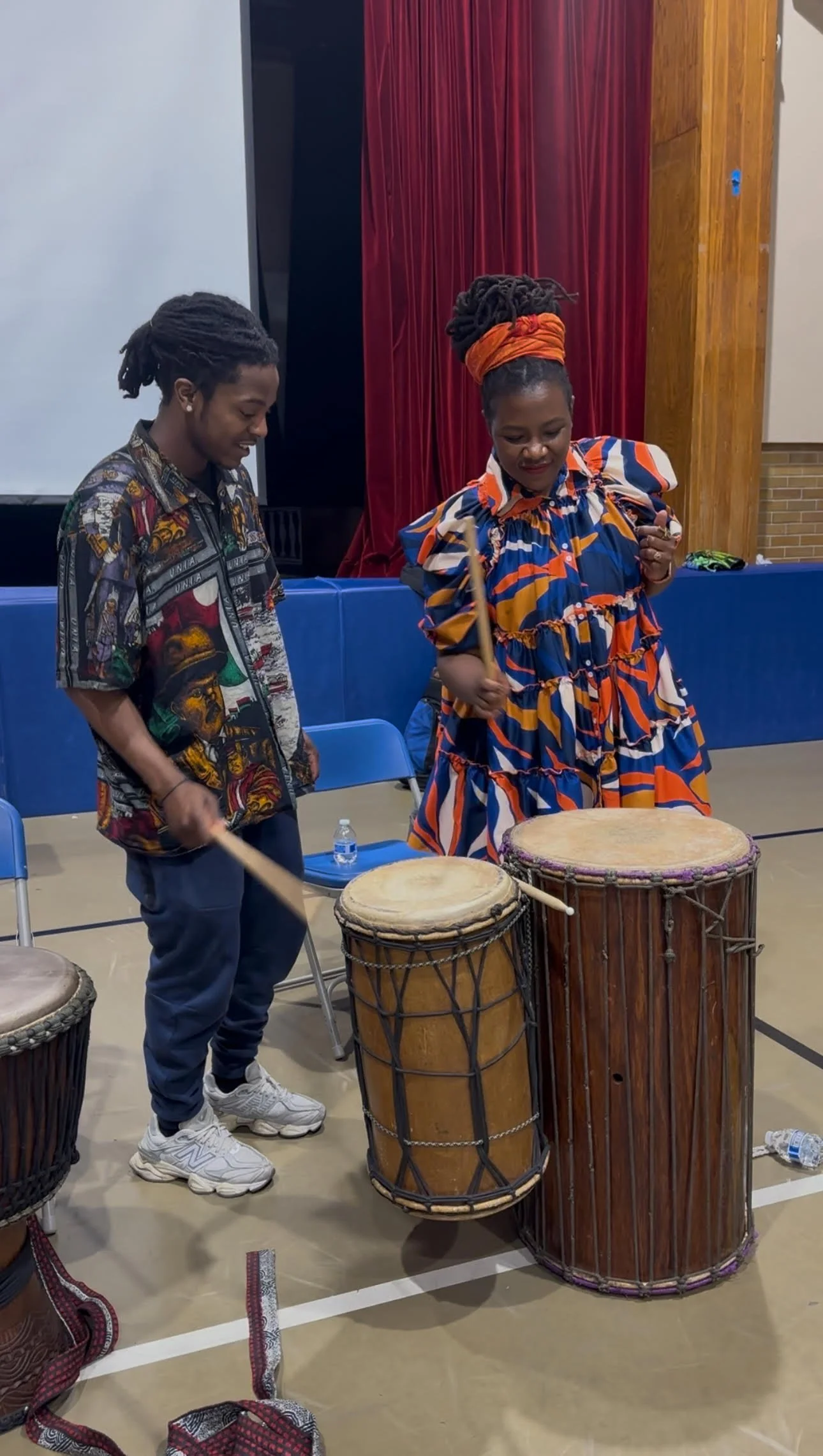 Two people playing African drums in a gymnasium, one woman with an orange headband and a colorful dress, and one man in a printed shirt and pants.