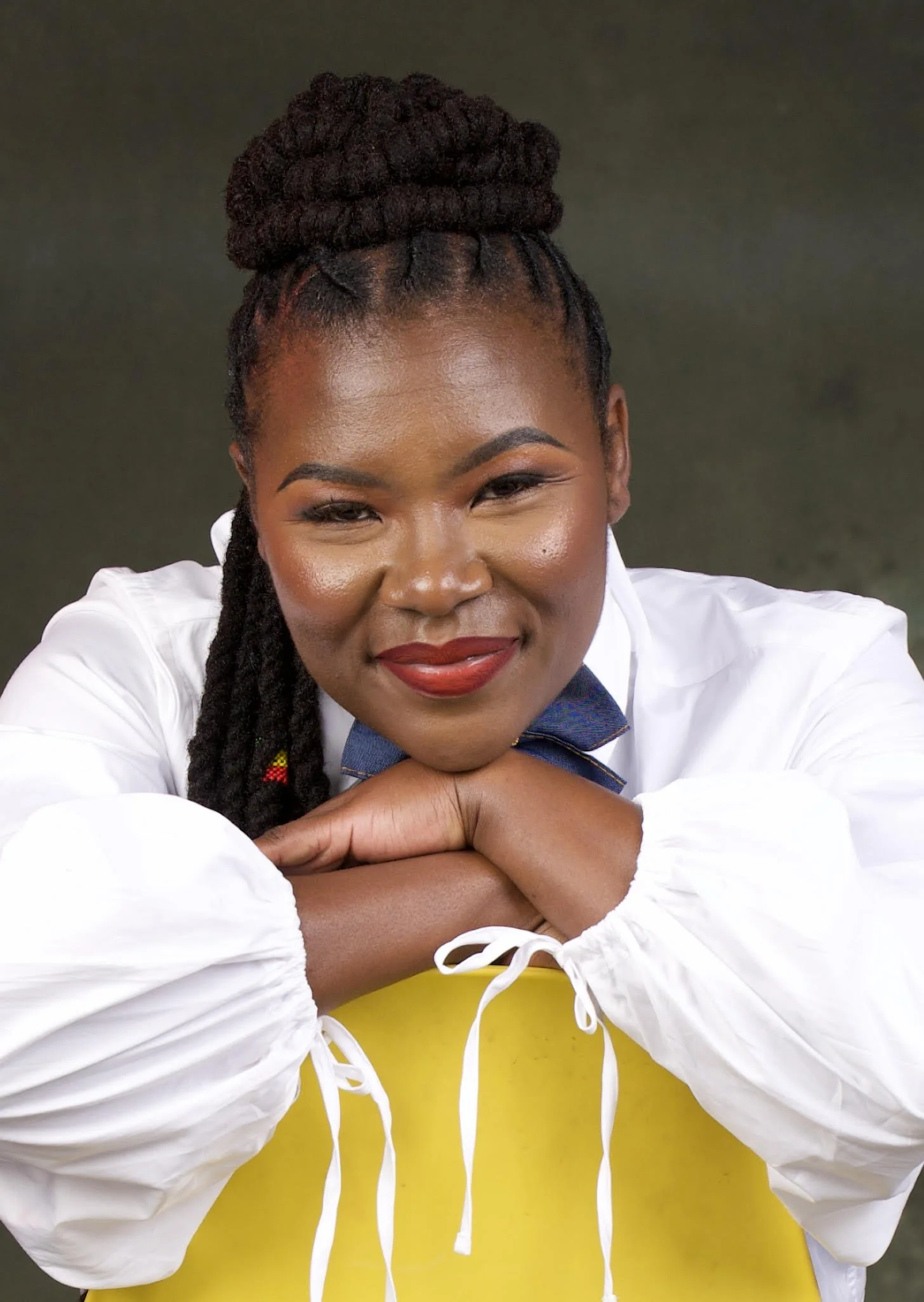 A woman with dark braided hair styled in an updo, wearing a white shirt and a blue bow tie, smiling with her chin resting on her crossed arms, in front of a yellow chair.