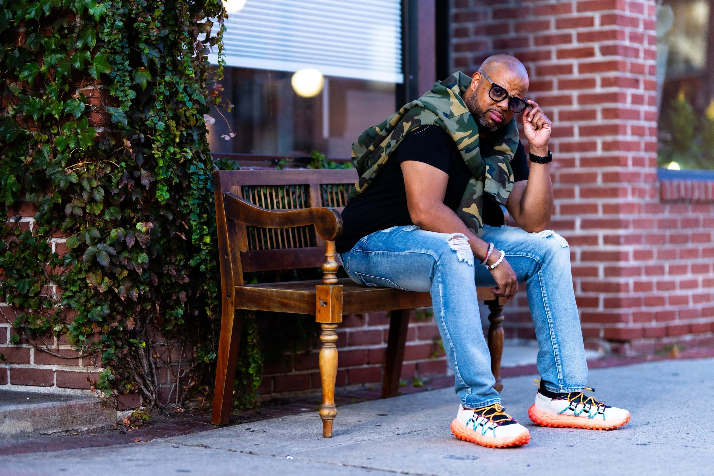 A man sitting on a park bench, wearing glasses, ripped jeans, colorful sneakers, and a camouflage jacket draped over his shoulders, with a brick building and ivy-covered wall in the background.