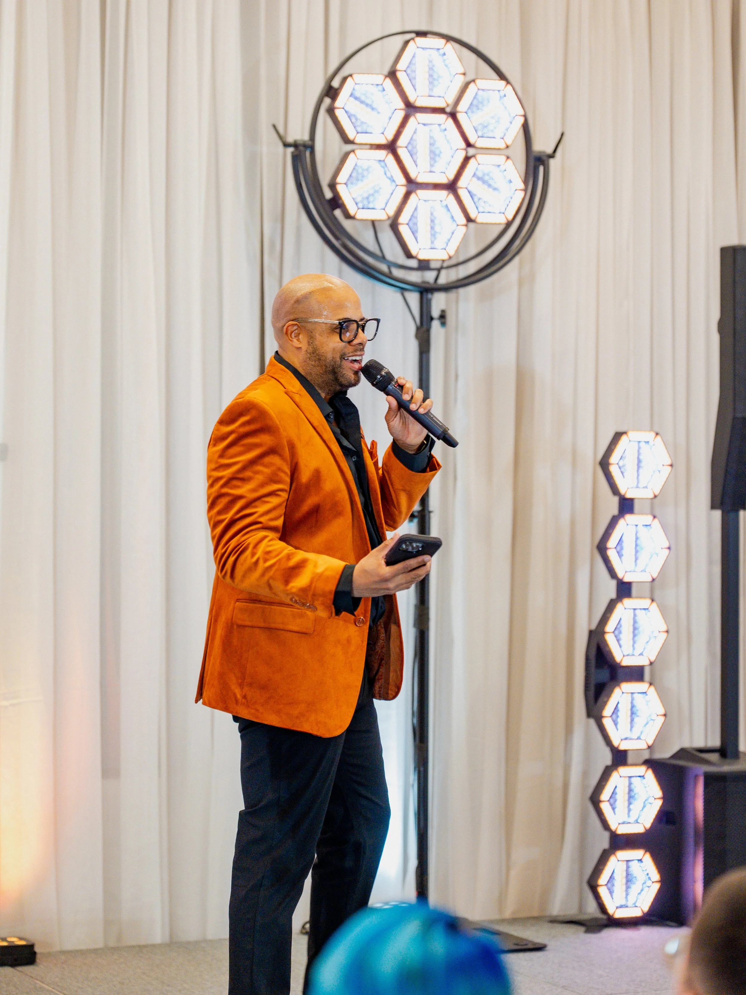 A man in an orange blazer and black glasses is speaking into a microphone at an indoor event, holding a phone in his other hand, with stage lighting and white curtains in the background.