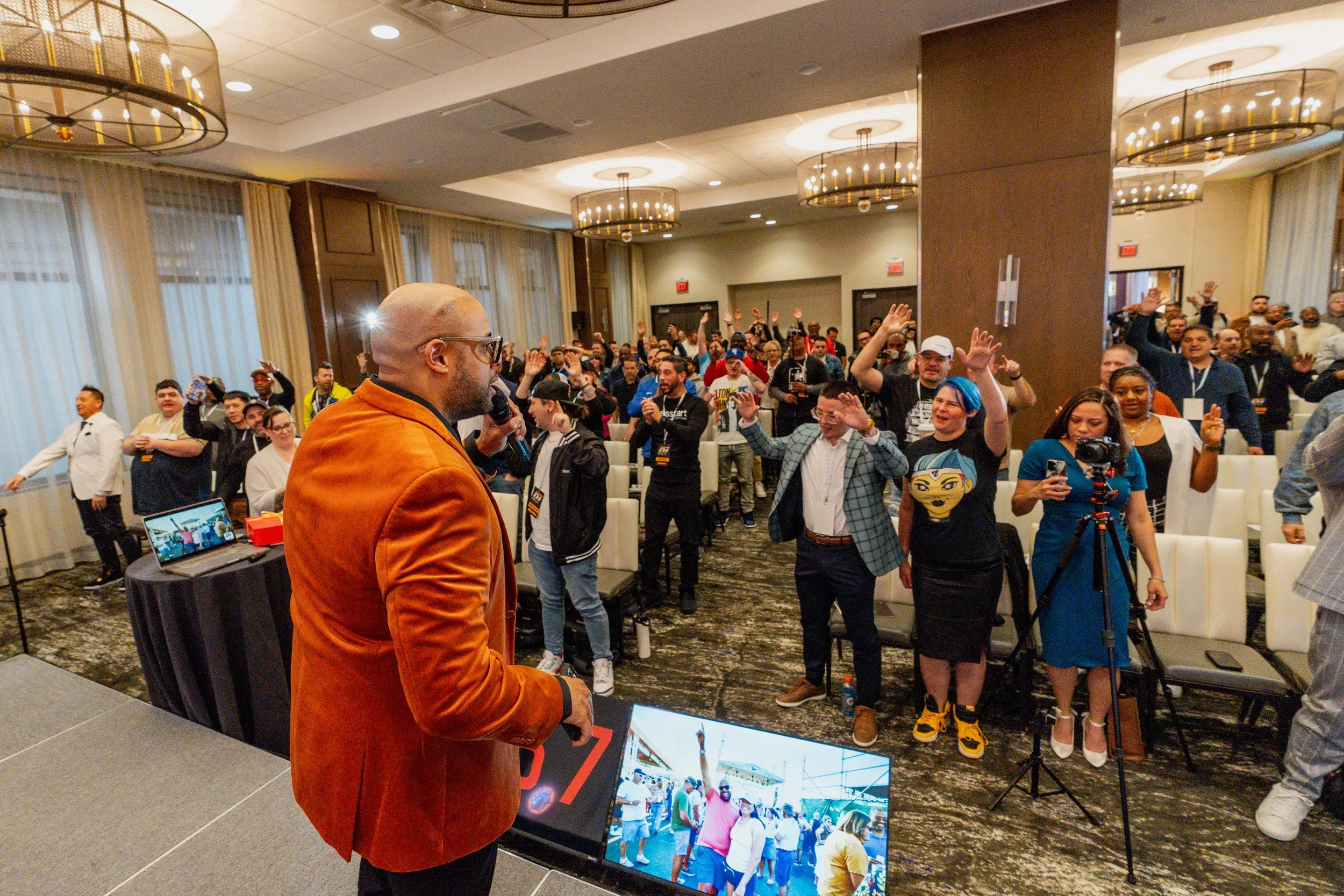 A speaker in an orange blazer is addressing a crowded audience in a conference room. The audience members are standing, some with raised hands, and appear engaged and happy. Several people are taking photos or videos, and there are cameras and a laptop on the stage in front of the speaker.