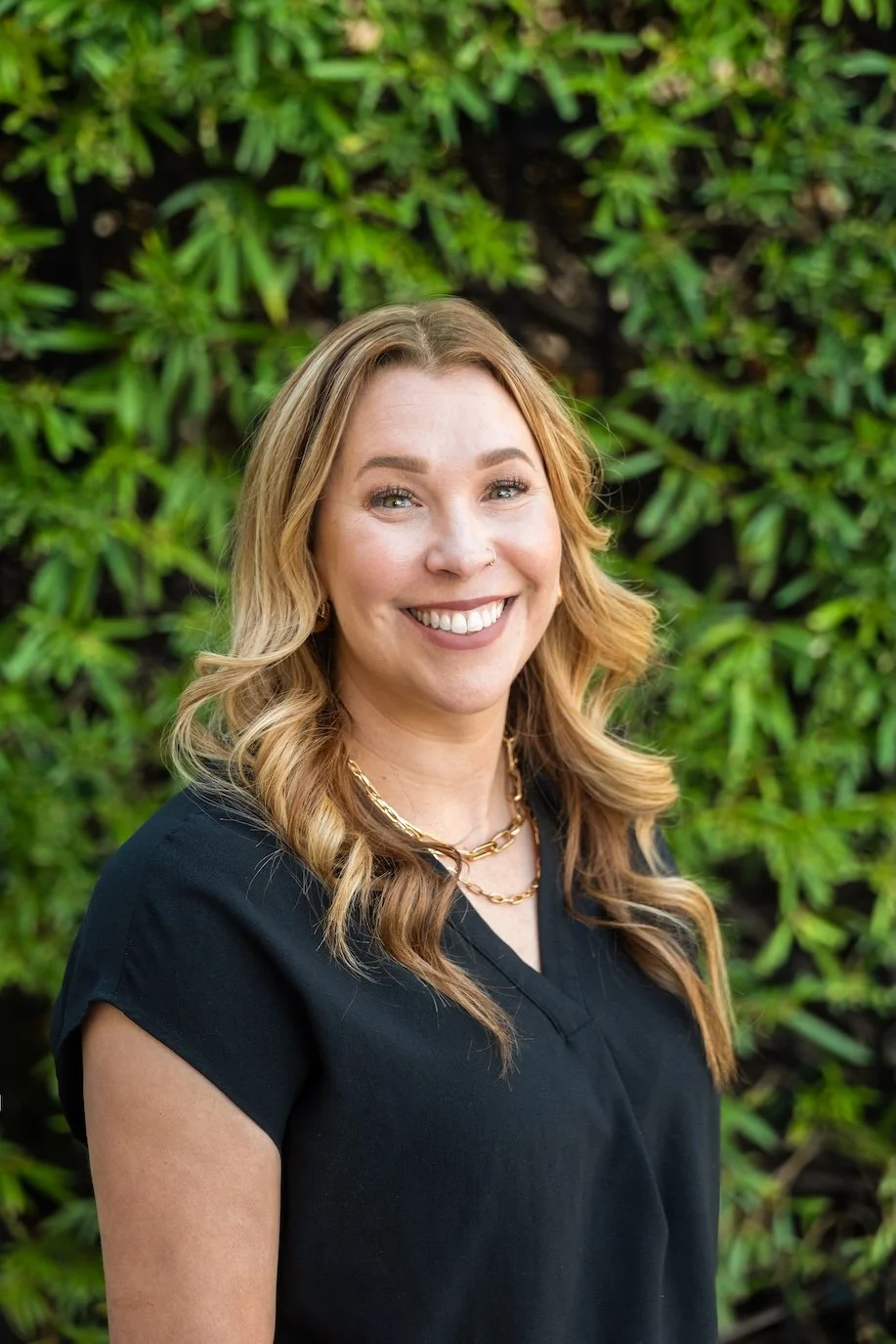 A woman with long wavy blonde hair, smiling with white teeth, wearing a black top and gold jewelry, standing outdoors in front of green leafy foliage.