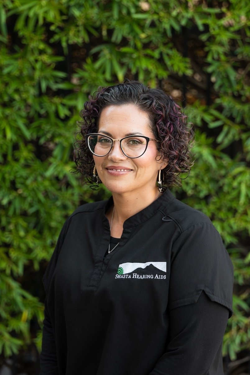 A woman with curly dark hair, glasses, and earrings, wearing a black shirt with a logo that reads 'Shasta Hearing Aids,' standing outdoors in front of green foliage.