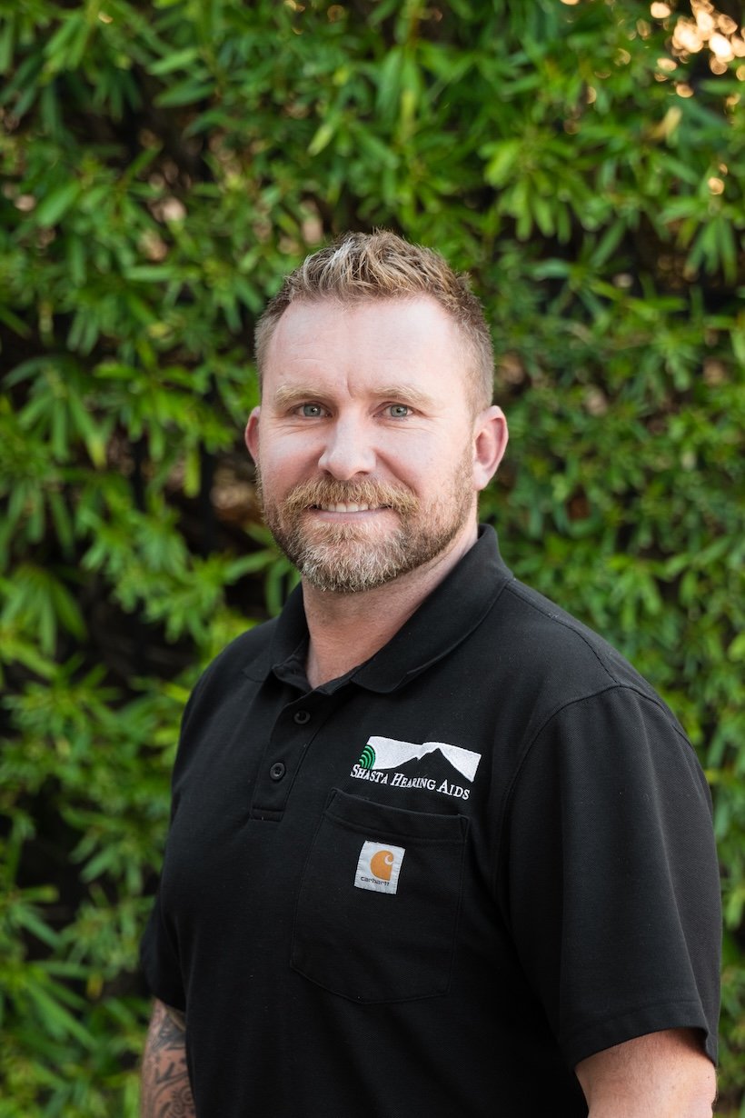A man with a beard and short hair, smiling, wearing a black polo shirt with a logo for Shasta Erawing AIDS and Carhartt, standing outdoors in front of green foliage.
