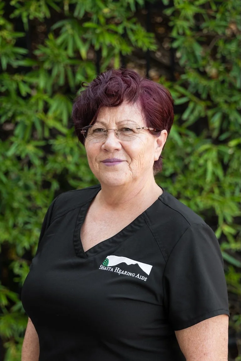 A woman with short, dark red hair and glasses, wearing a black shirt with a logo that says "Shasta Hearing Aids," standing outdoors in front of green leafy bushes.