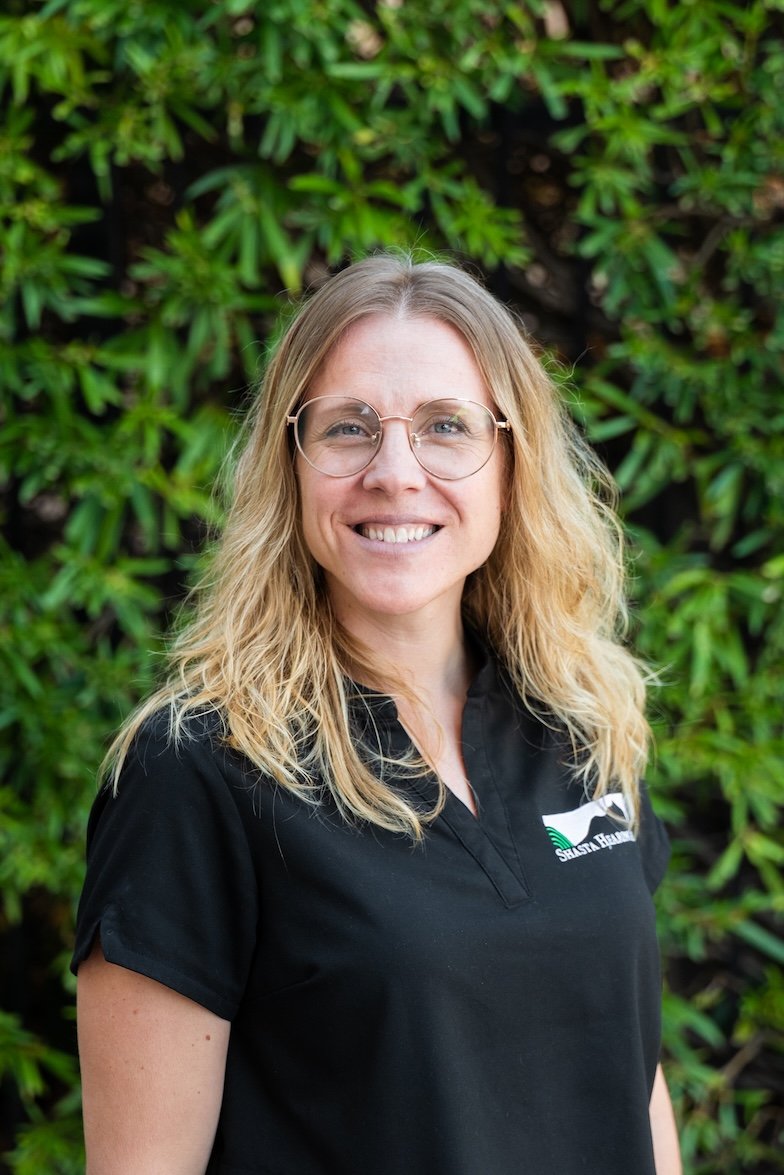 A woman with long blonde hair and glasses smiling outdoors in front of green foliage, wearing a black polo shirt with a logo.