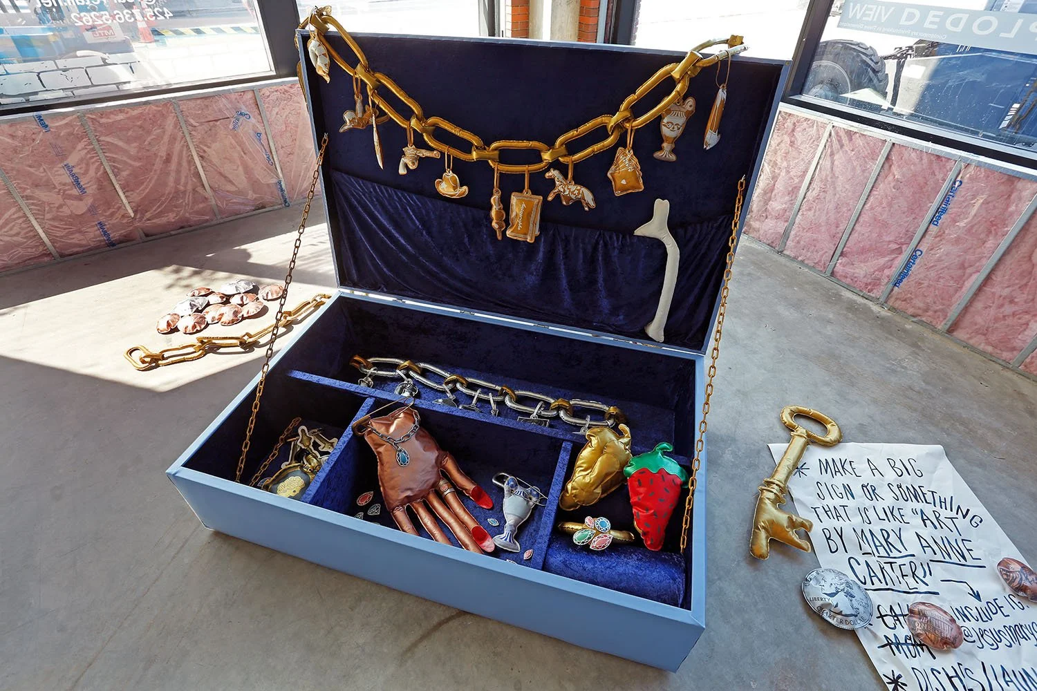 Open 8 foot tall jewelry box with gold and silver chains, charm pendants, and jewelry accessories inside and outside, displayed in a brightly lit space with construction or renovation materials on the walls.