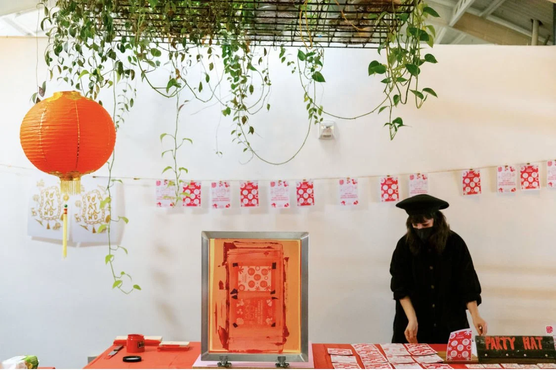 A person dressed in black with a black hat and face mask live screen printing at a wedding decorated with red hanging lanterns, ivy, and postcard prints drying on a string against a white wall.