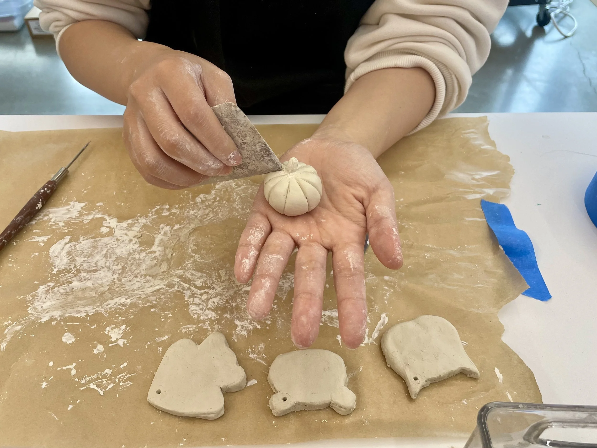Person making a ceramic pumpkin sculpture, carving lines with a tool. Several unglazed ceramic ornaments, including a heart, a pumpkin, and a ghost, are on a piece of paper on the table.