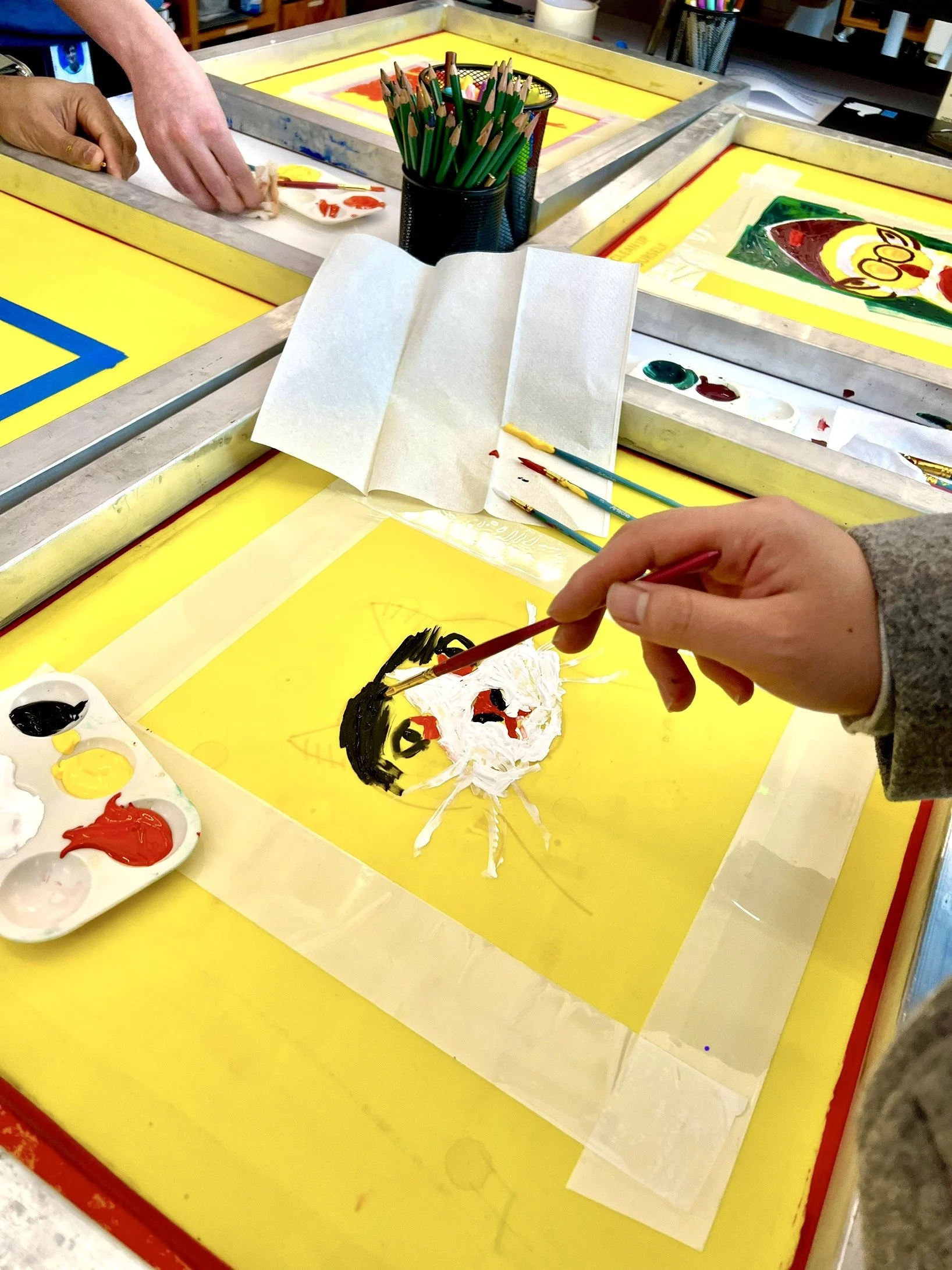 Person creating a pet portrait painting of a cat using white, yellow, black, and red paint on a yellow silk screen at an art table with other art supplies nearby.
