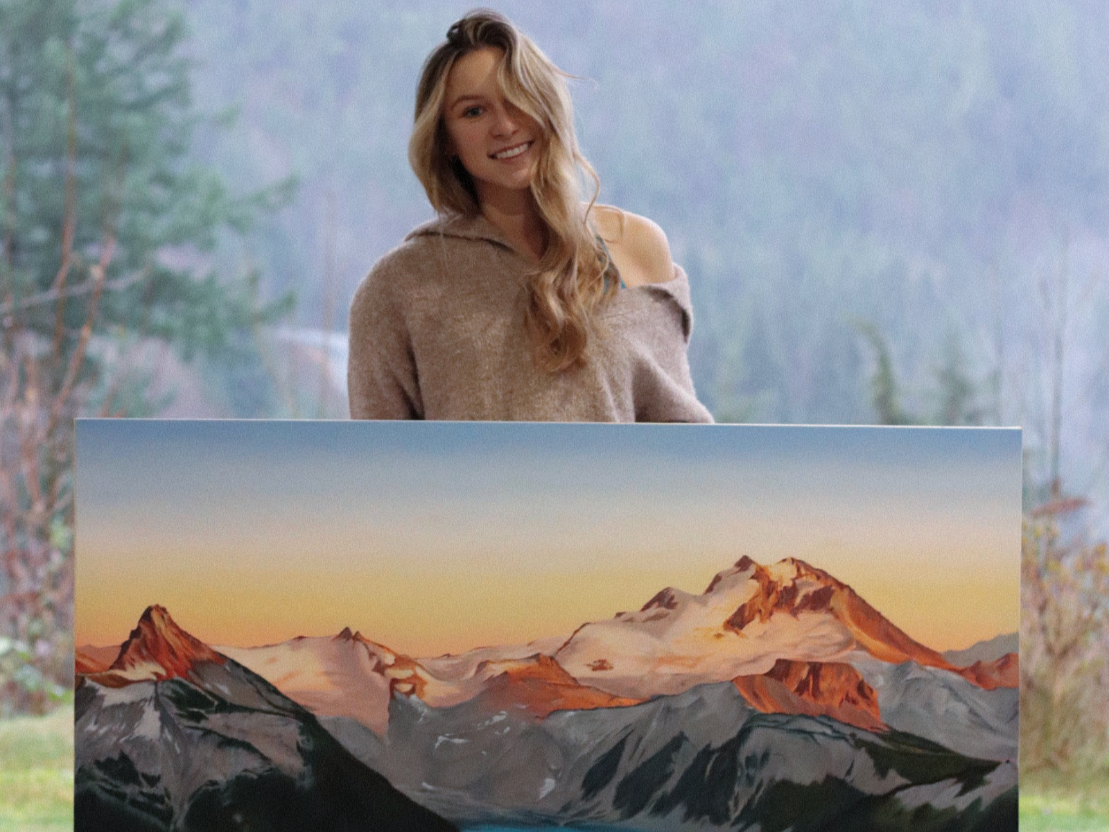 A woman with long wavy blonde hair wearing a beige off-shoulder sweater standing outdoors with a mountain painting or poster in front of her.