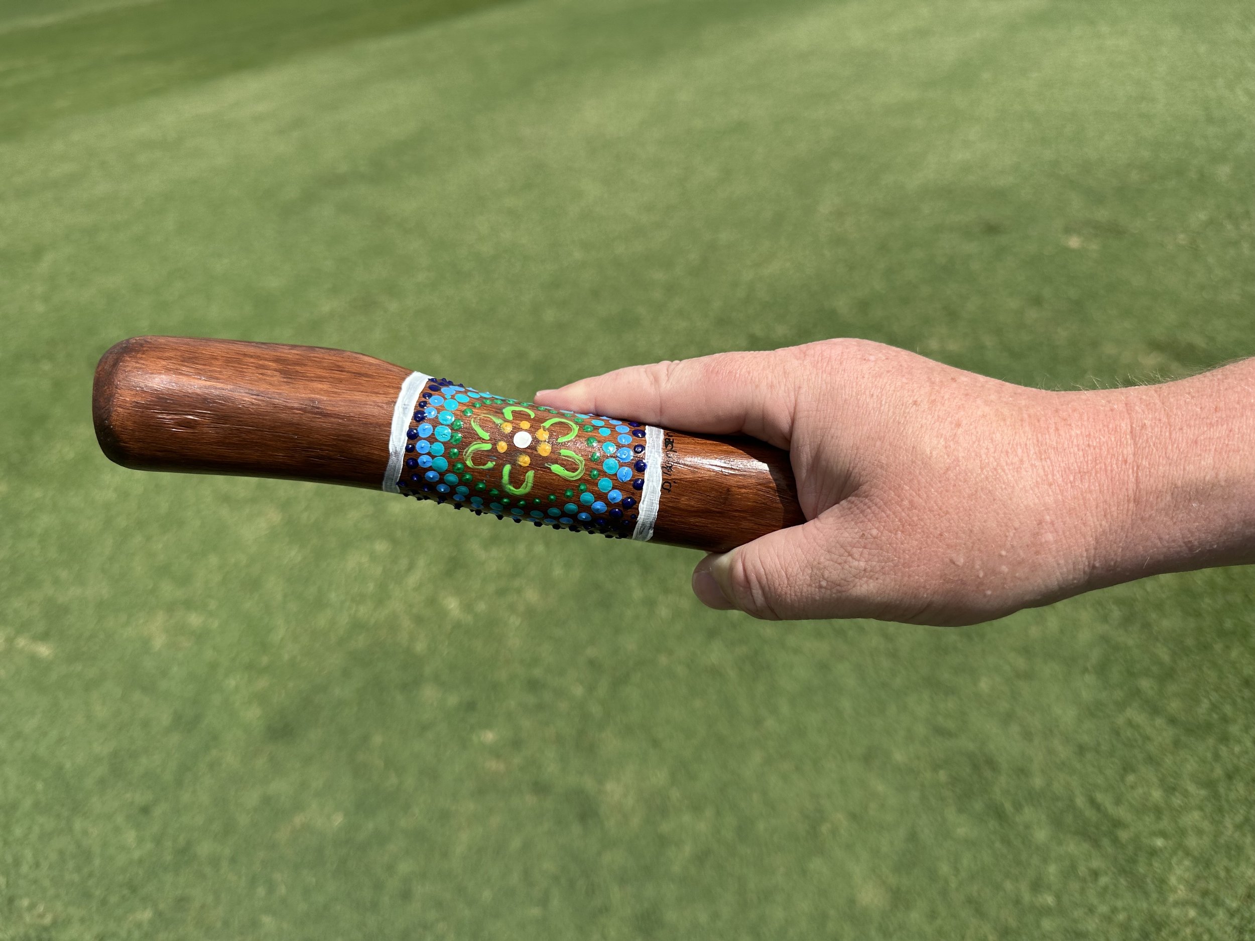 A hand holding a wooden yarning stick with a decorated grip, against a background of green grass.