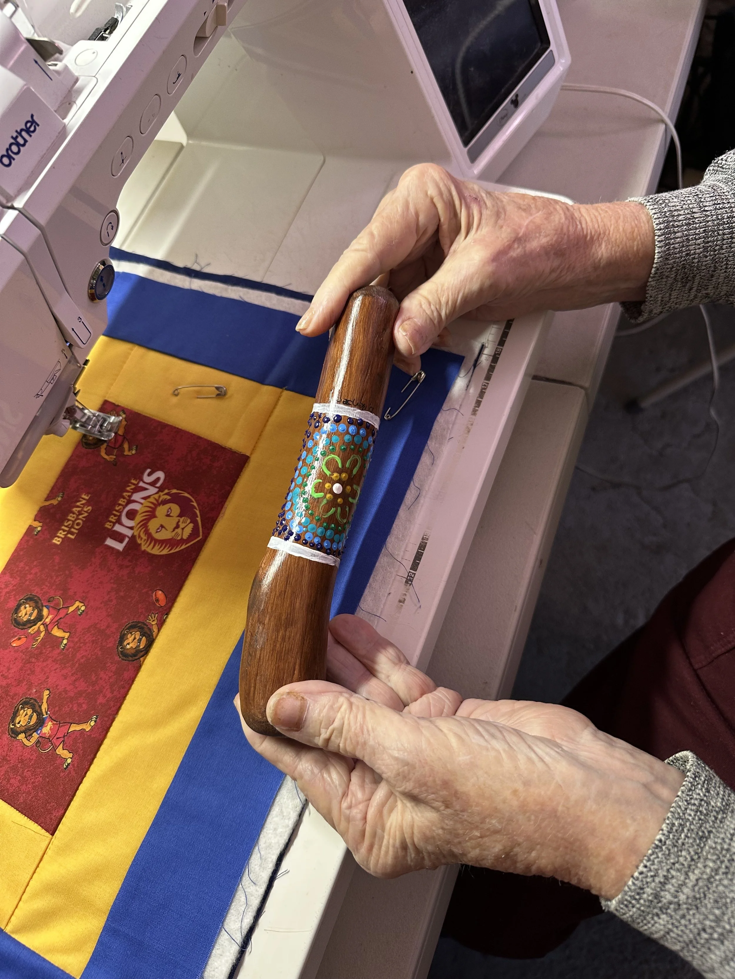 A person holding a decorated wooden yarning stick, possibly for crafting or sewing, in front of a sewing machine on a table with yellow, blue, and red fabric featuring lions and the word 'LIONS'.
