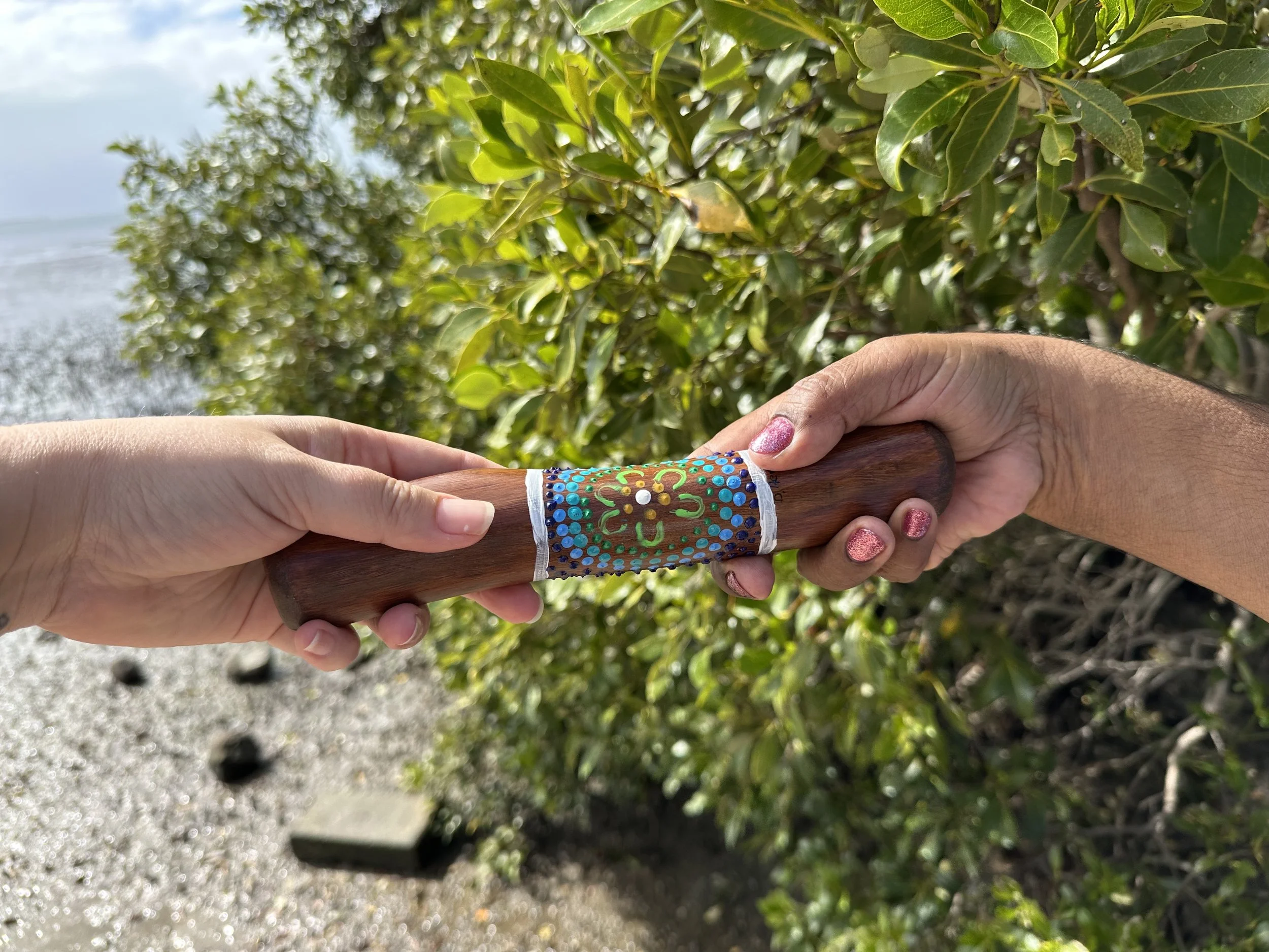 Two hands hold a decorated yarning stick together outdoors near greenery and water.
