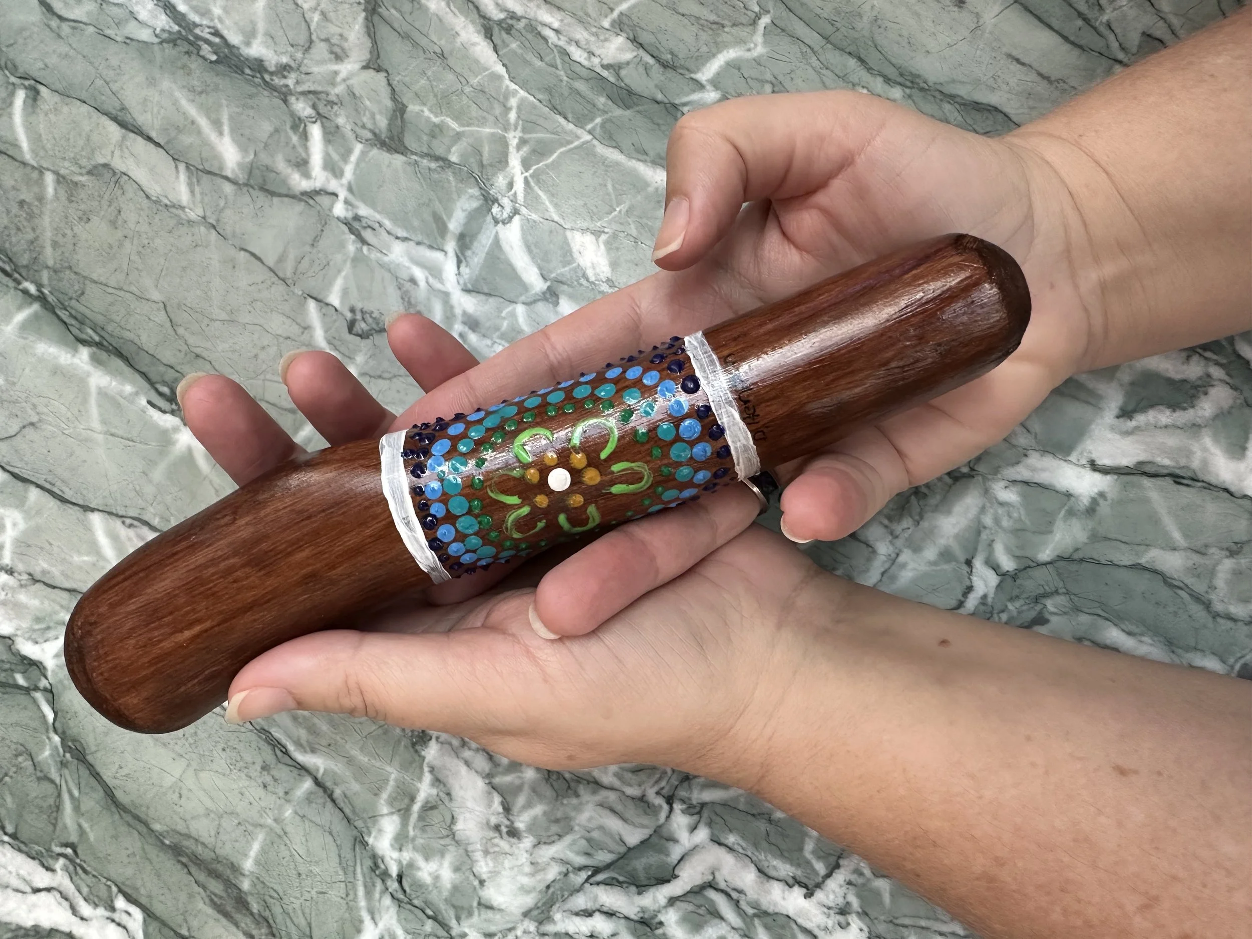 Person holding a decorated wooden yarning stick with painted dots and a floral pattern, against a gray marble background.