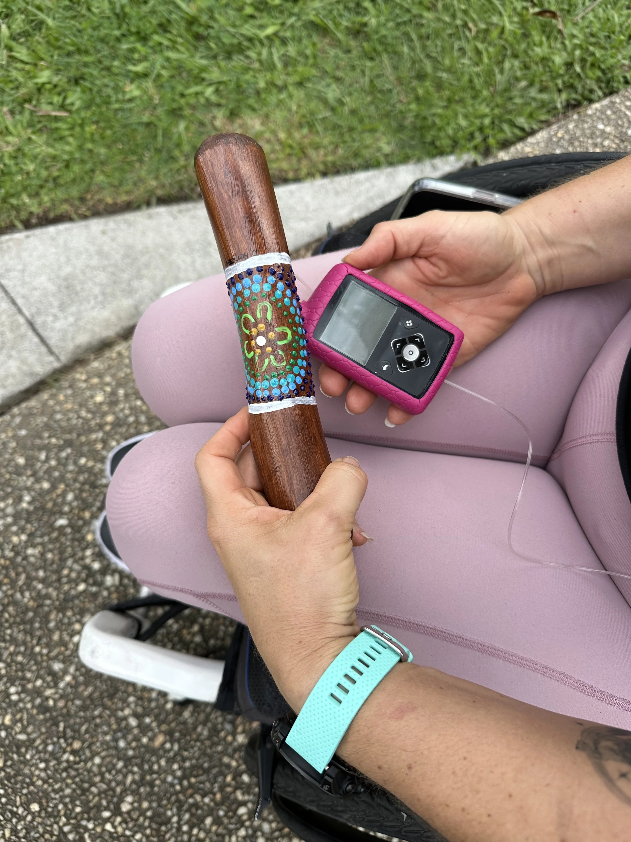 A person sitting outdoors holding a handmade wooden yarning stick with colorful dot patterns and a small electronic device with a pink case. The person is wearing pink pants and a turquoise watch, with green grass and a path in the background.