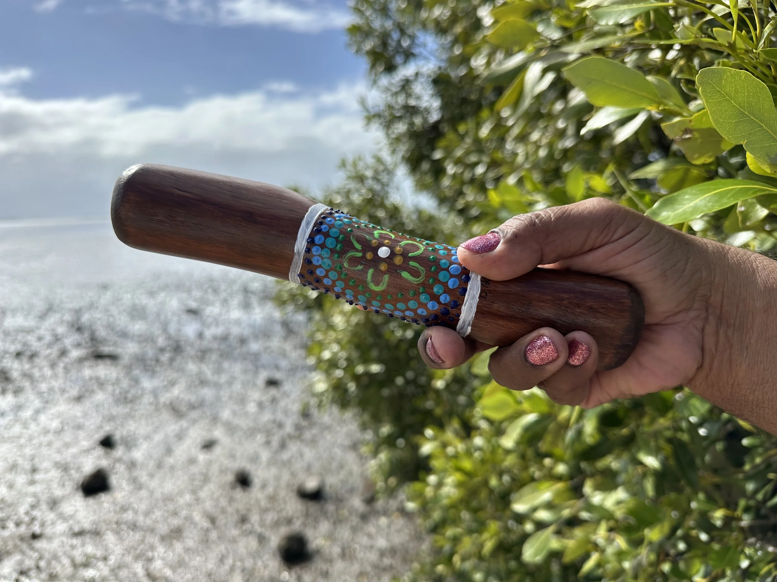 A person's hand holding a wooden yarning stick with a decorated band, on a background of green leaves, gray gravel, and blue sky with clouds.