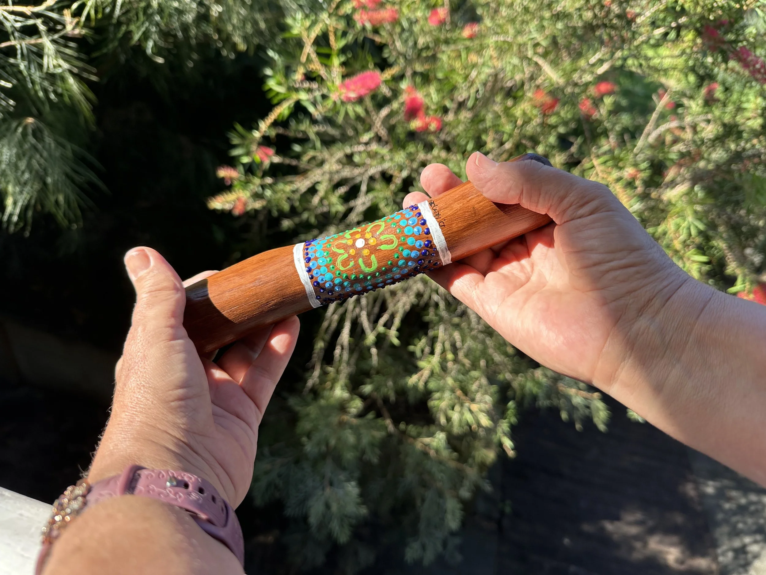 Person holding a decorated wooden stick with colorful dot and flower patterns painted on it, outdoors with green plants in the background.