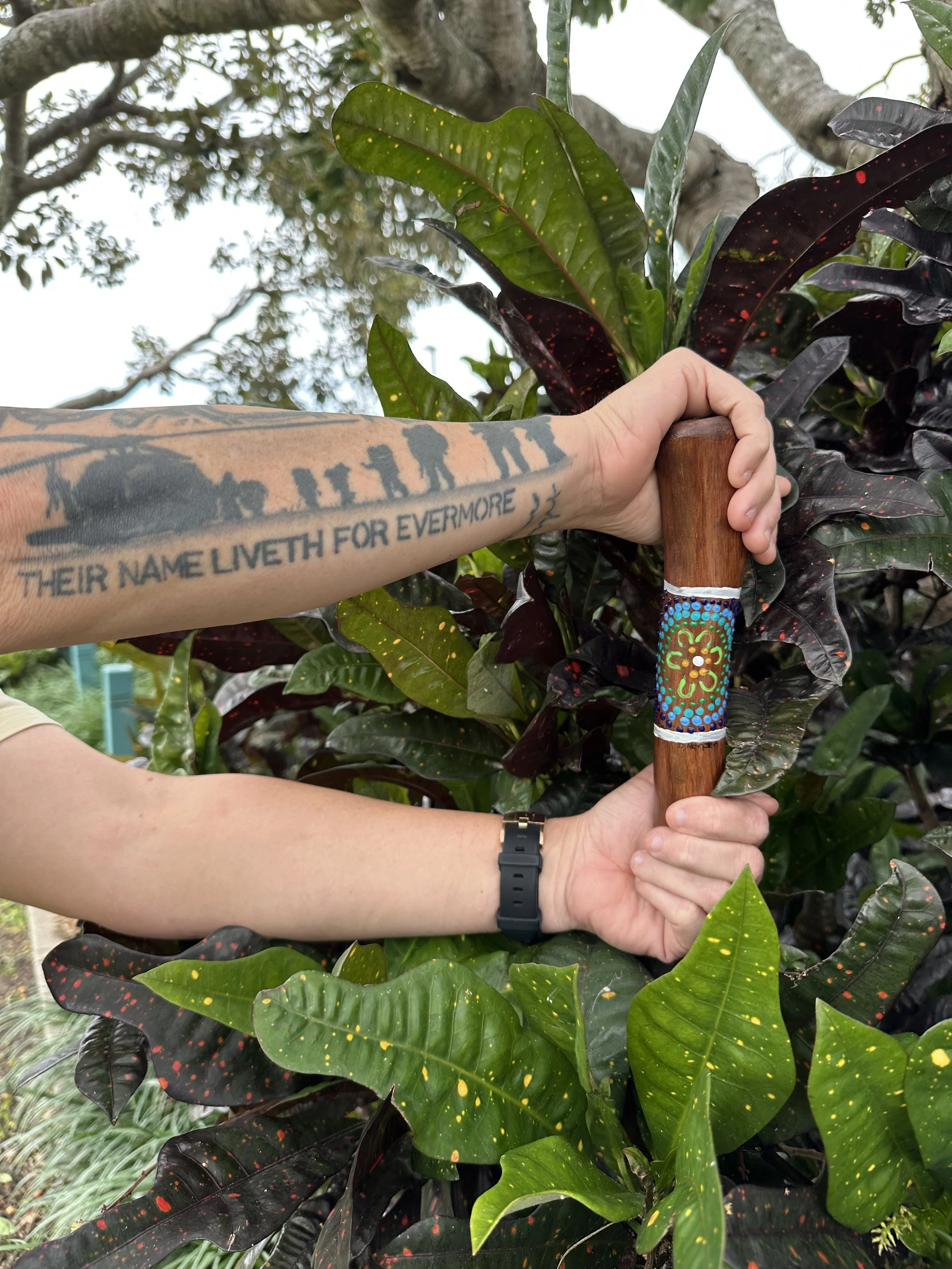 A person with tattoos and a watch holding a wooden yarning stick with colorful, painted designs among dark green and purple leaves.