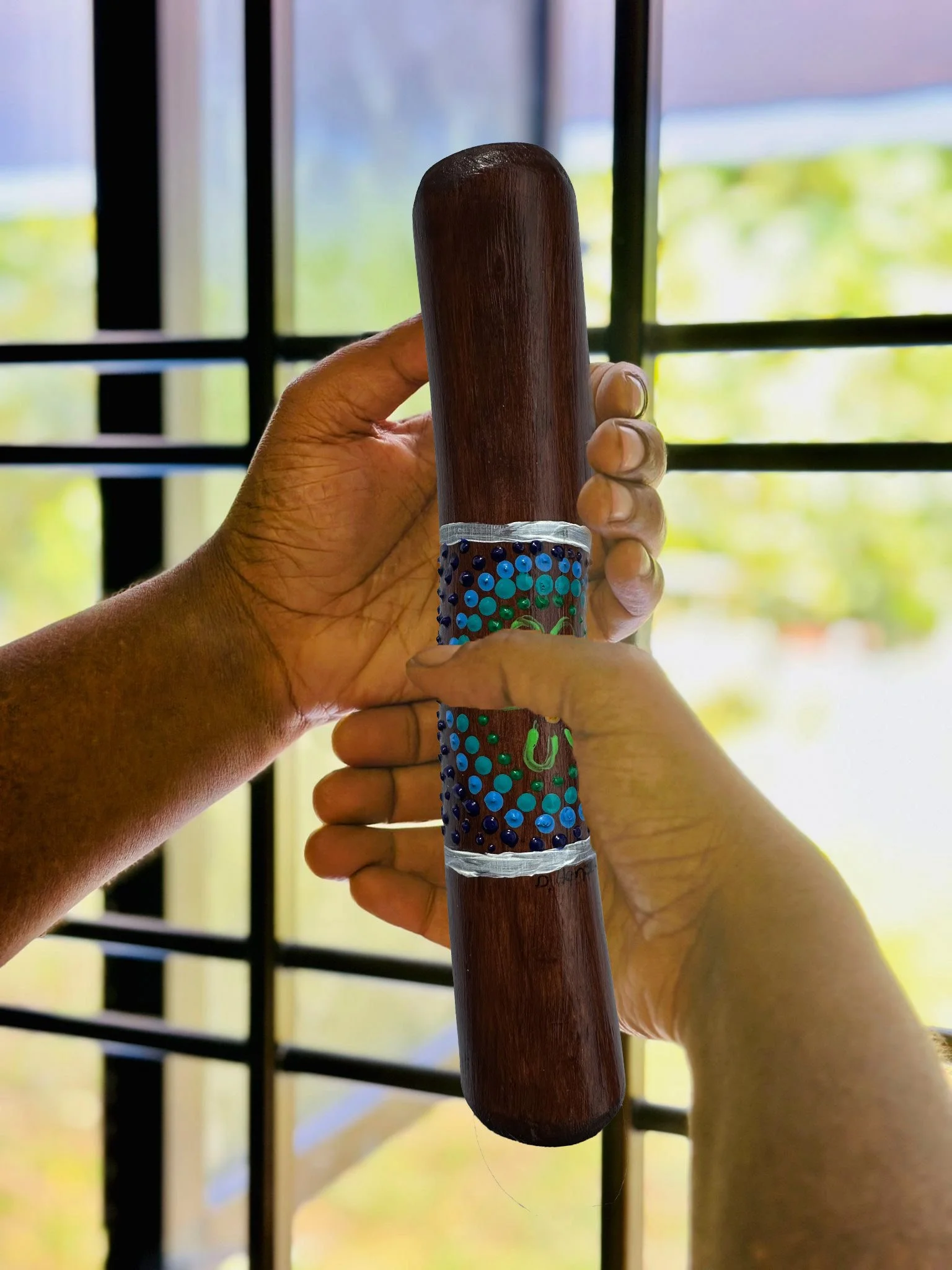 Hands holding a decorated wooden yarning stick with colorful dots and a small U shape design, in front of a window with a grid pattern.