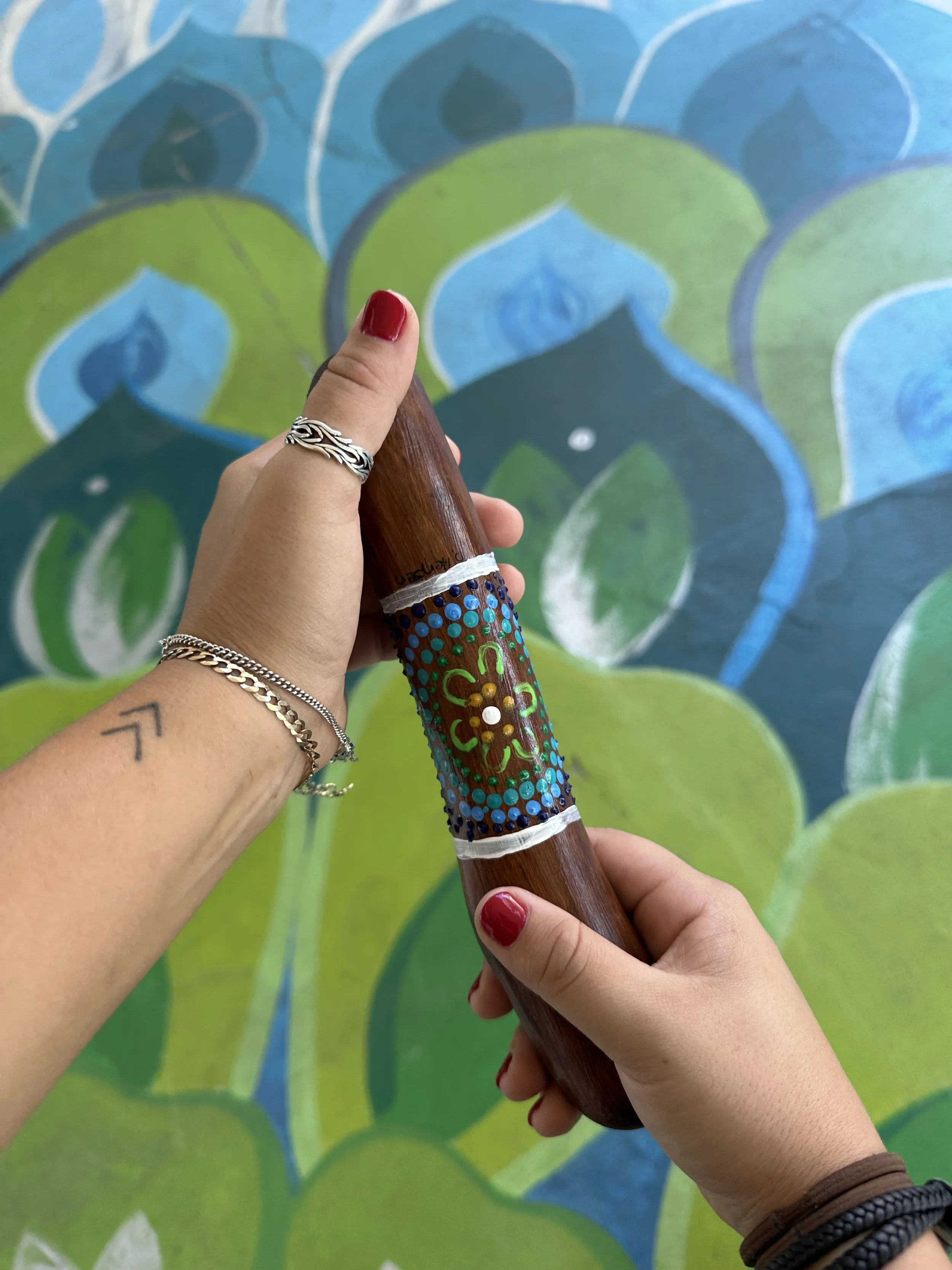 A person holding a decorated wooden yarning stick with colorful dot and floral patterns painted on a band around the middle. The person has red-painted nails, jewelry, and tattoos on their arm, against a vibrant abstract blue and green background.