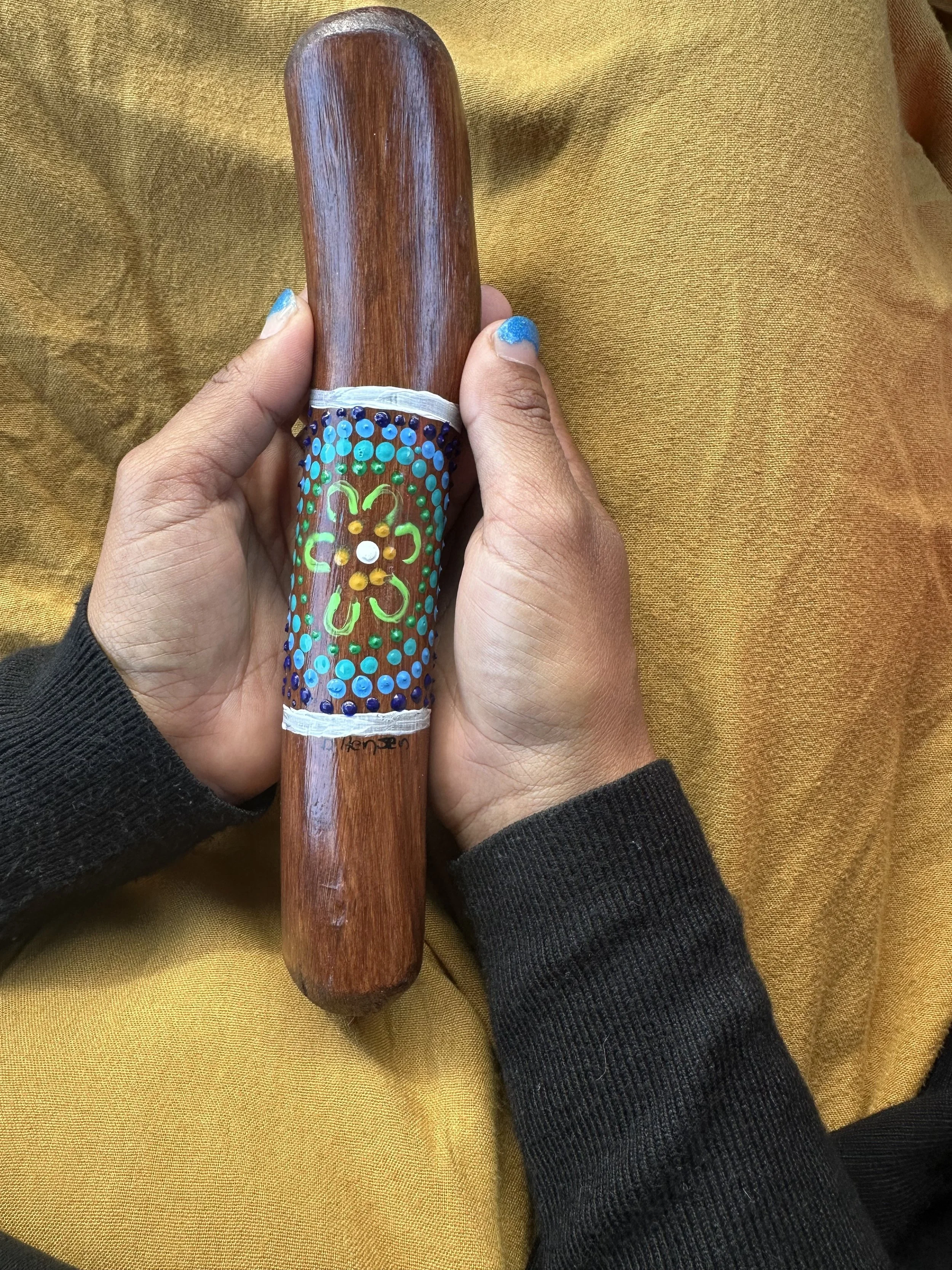 A hand holding a wooden yarning stick with painted and dotted designs, resting on a yellow fabric background.