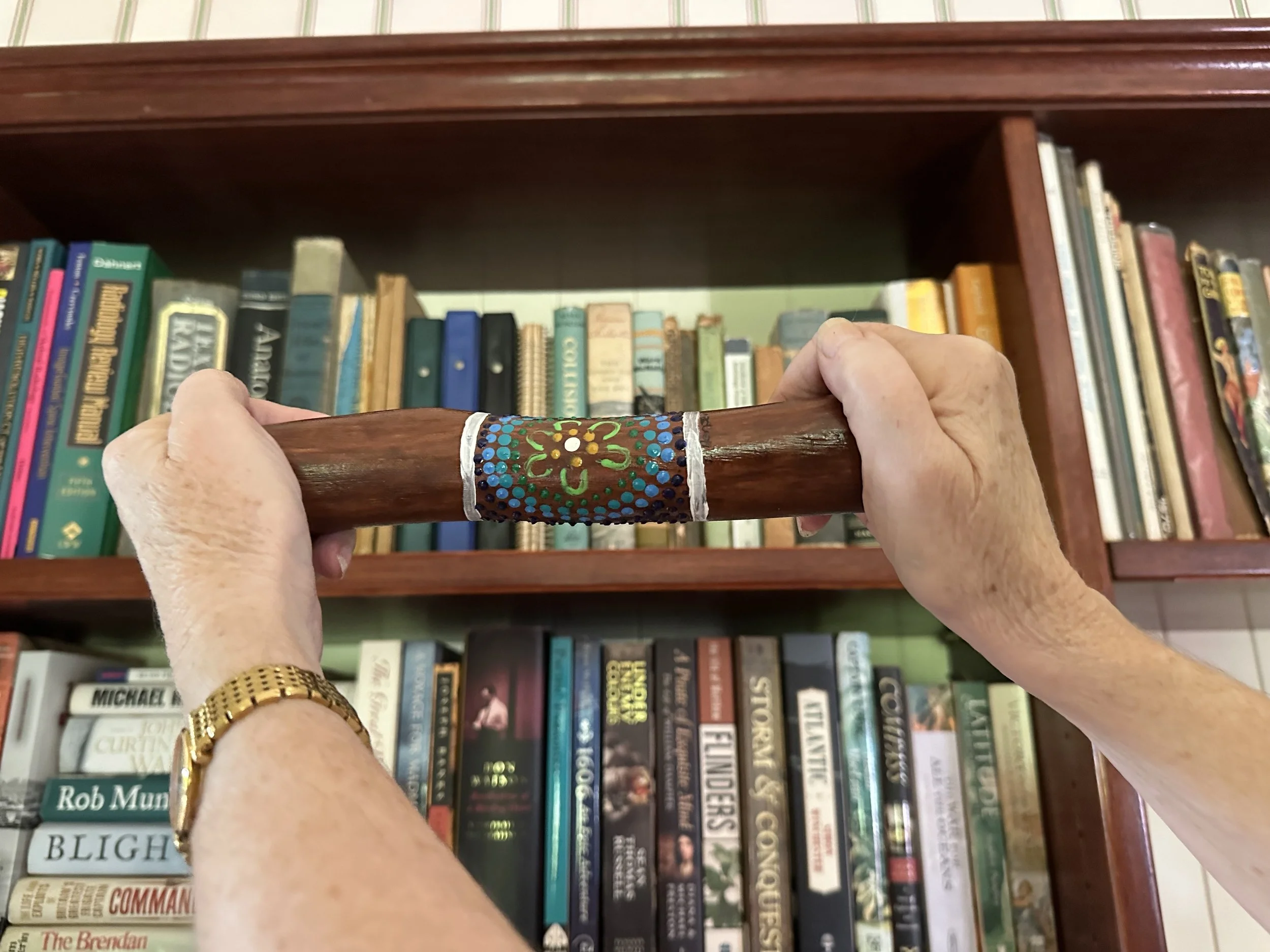 Person holding a decorated wooden yarning stick in front of a bookshelf filled with books.