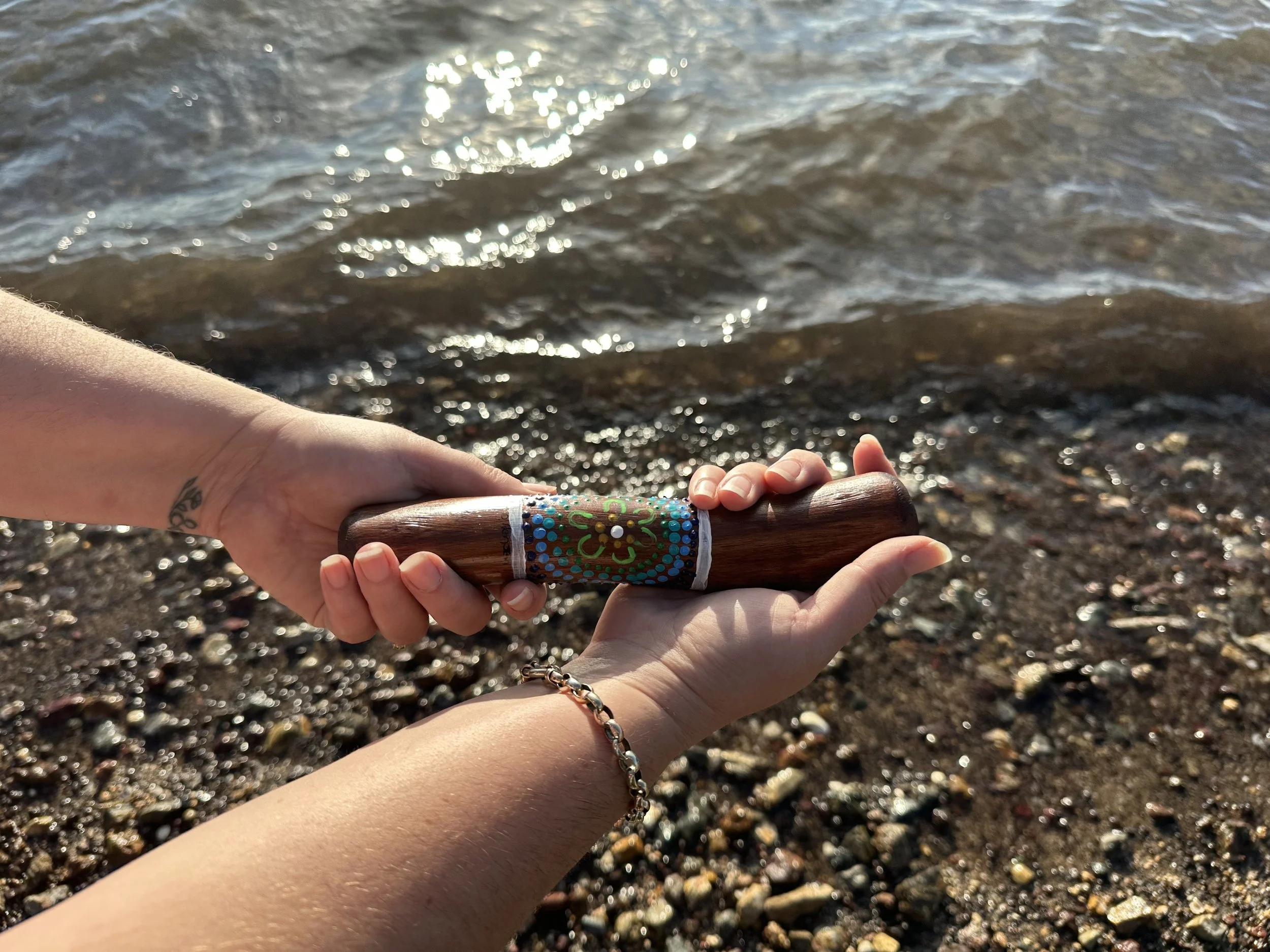 Two hands, one holding a wooden yarning stick and the other supporting it, near a body of water with a pebbled shoreline.