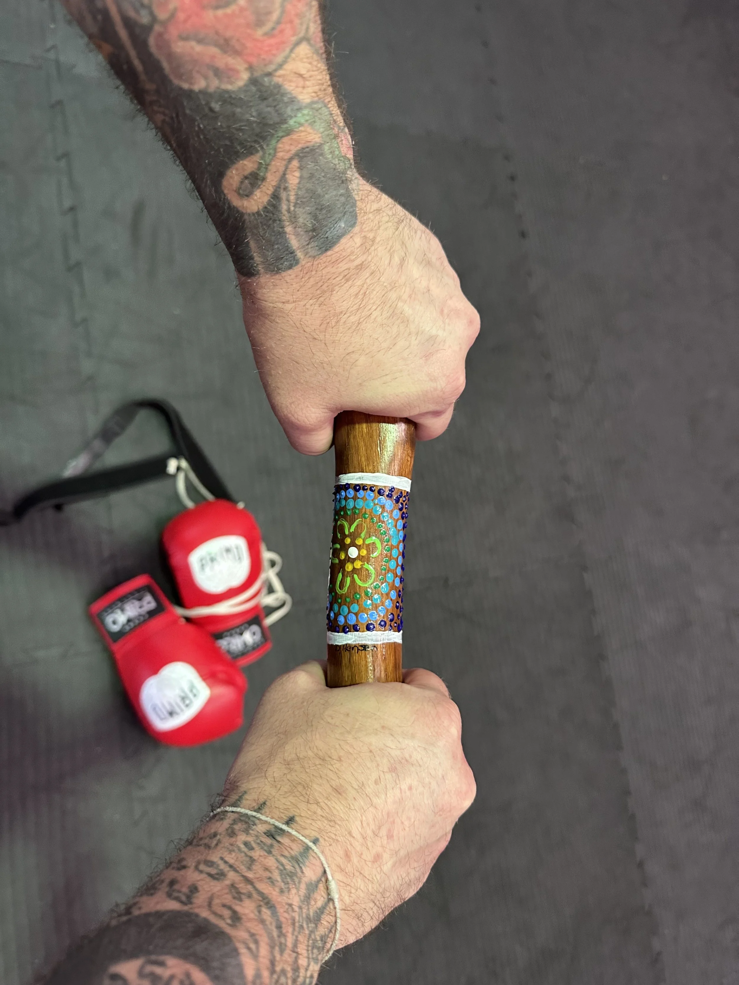 A person with tattoos on both arms holding a small wooden yarning stick with decorative colorful dots and a floral design on it. Red boxing gloves are seen on the floor nearby.