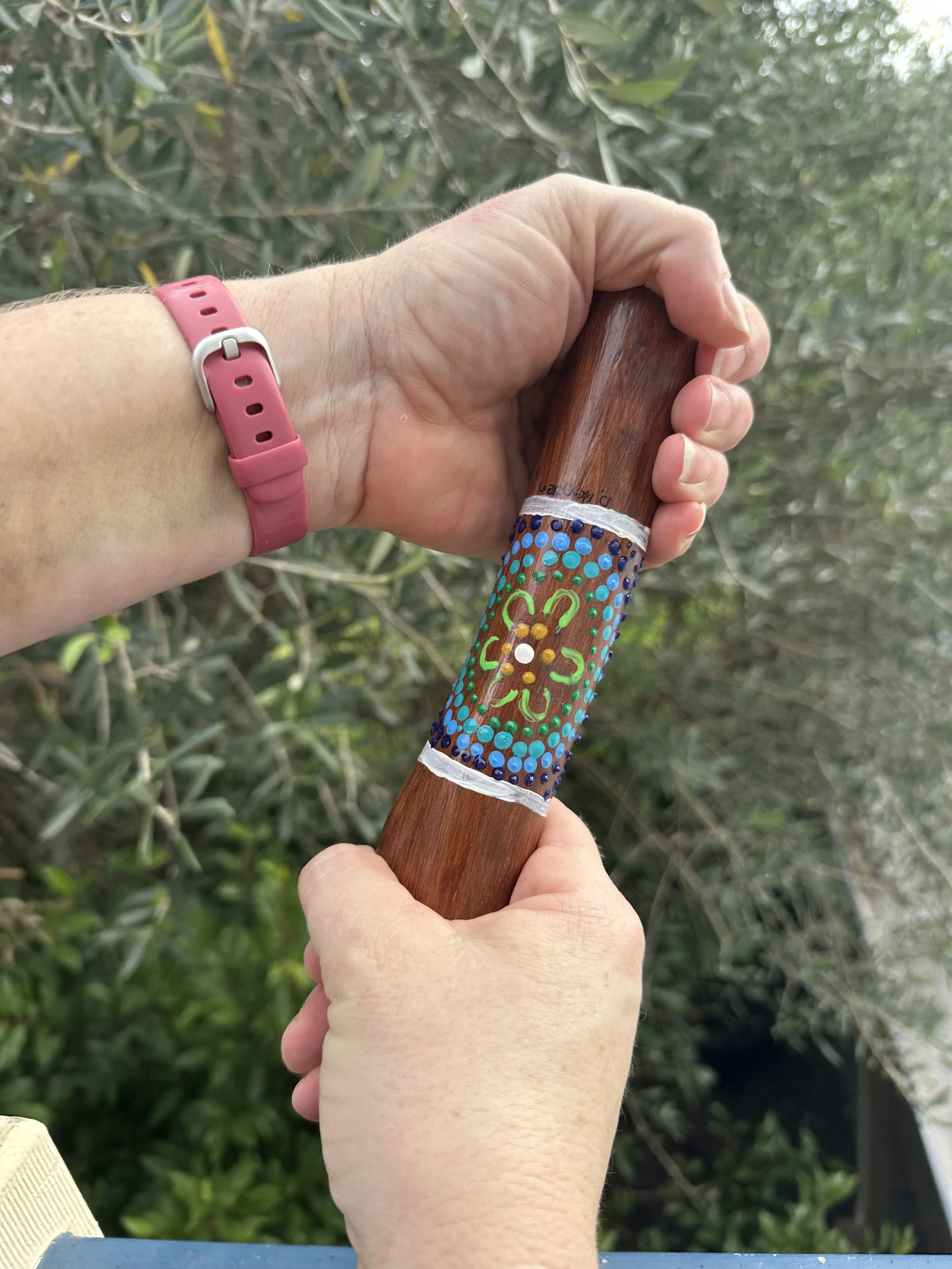 Person holding a wooden yarning stick with a colorful decorated band in front of green foliage.