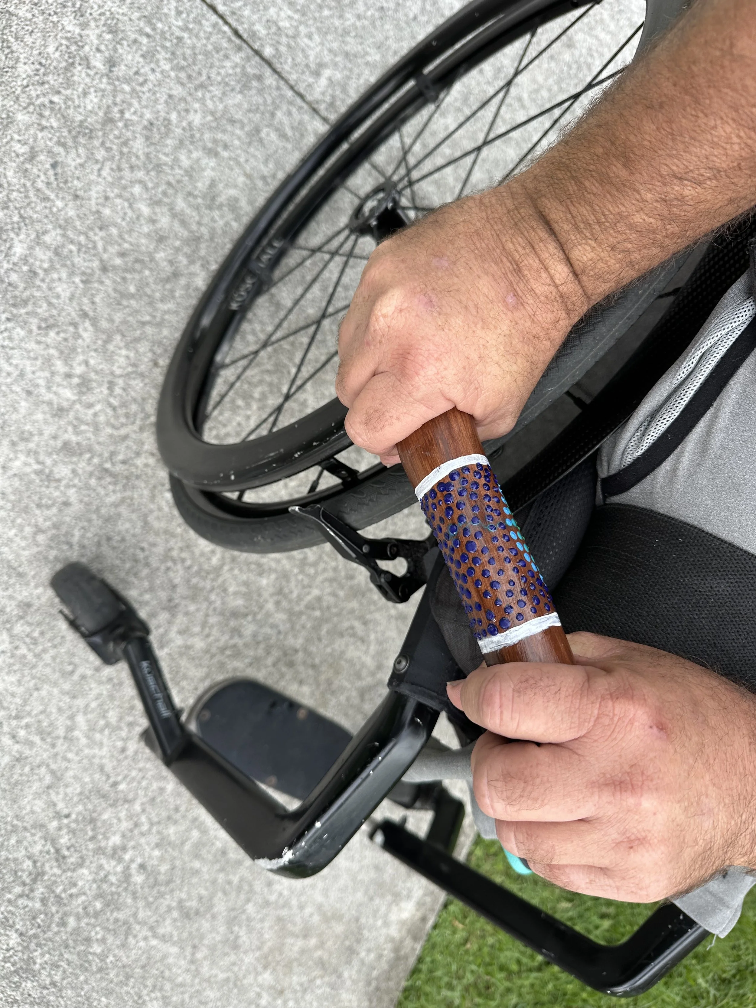 Close-up of a person holding a yarning stick with a wooden and blue patterned grip on a wheelchair.